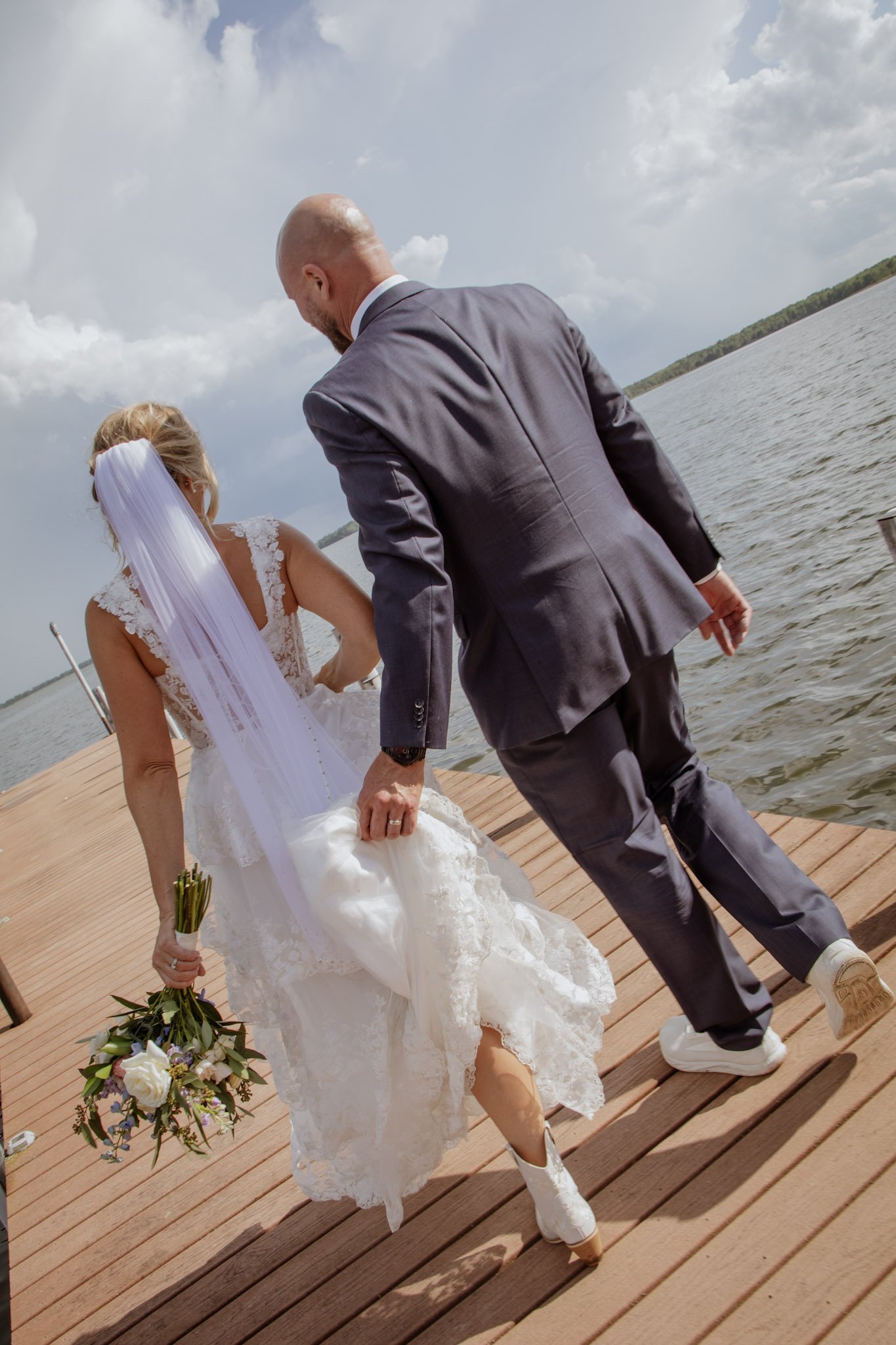 A bride and groom standing on a wooden dock near a body of water, holding hands, with the bride holding a bouquet of flowers, during a wedding ceremony outdoors on a cloudy day.