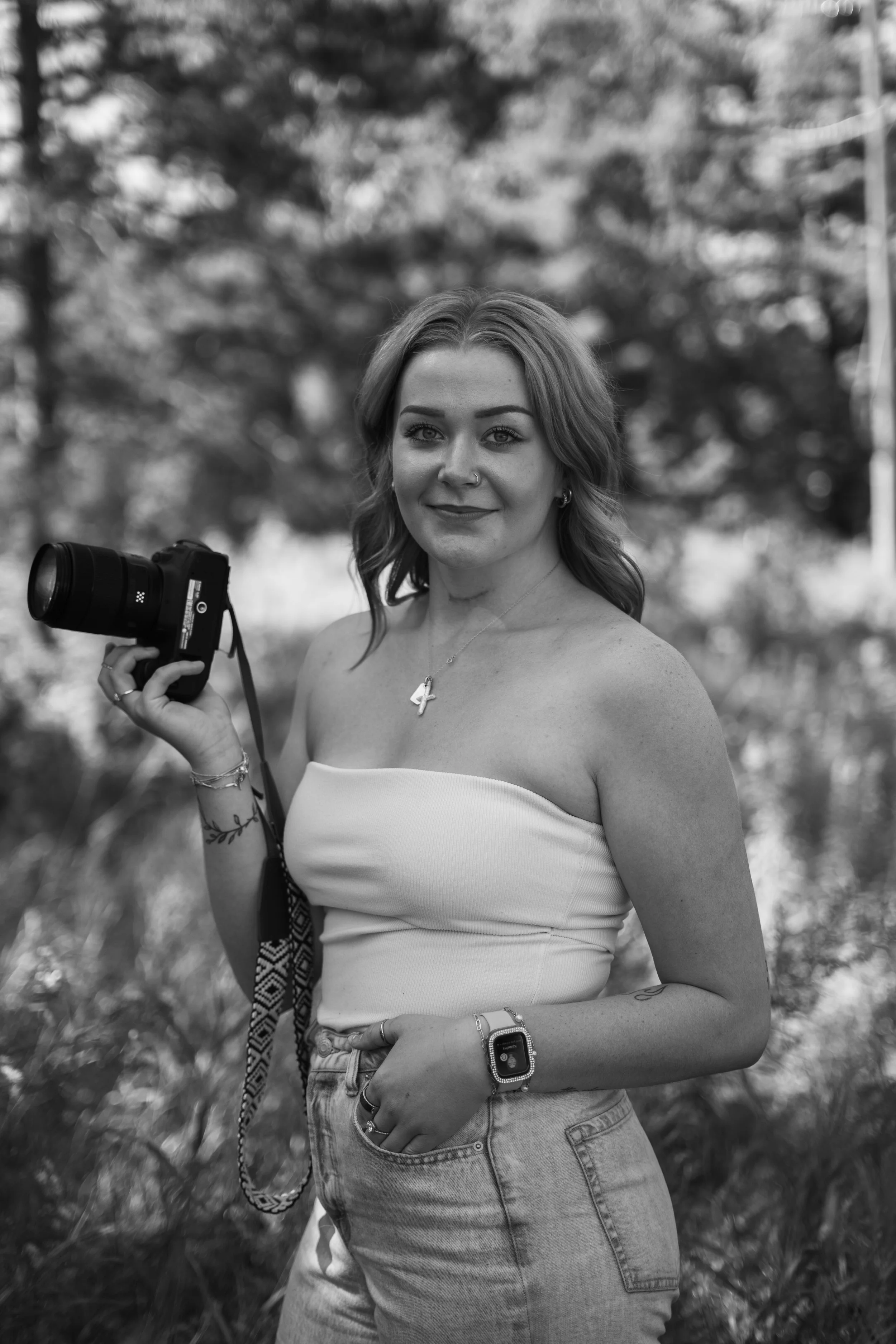 A young woman outdoors holding a camera, wearing a strapless top and jeans, with trees in the background. Minnesota Photographer. Travel Photographer.