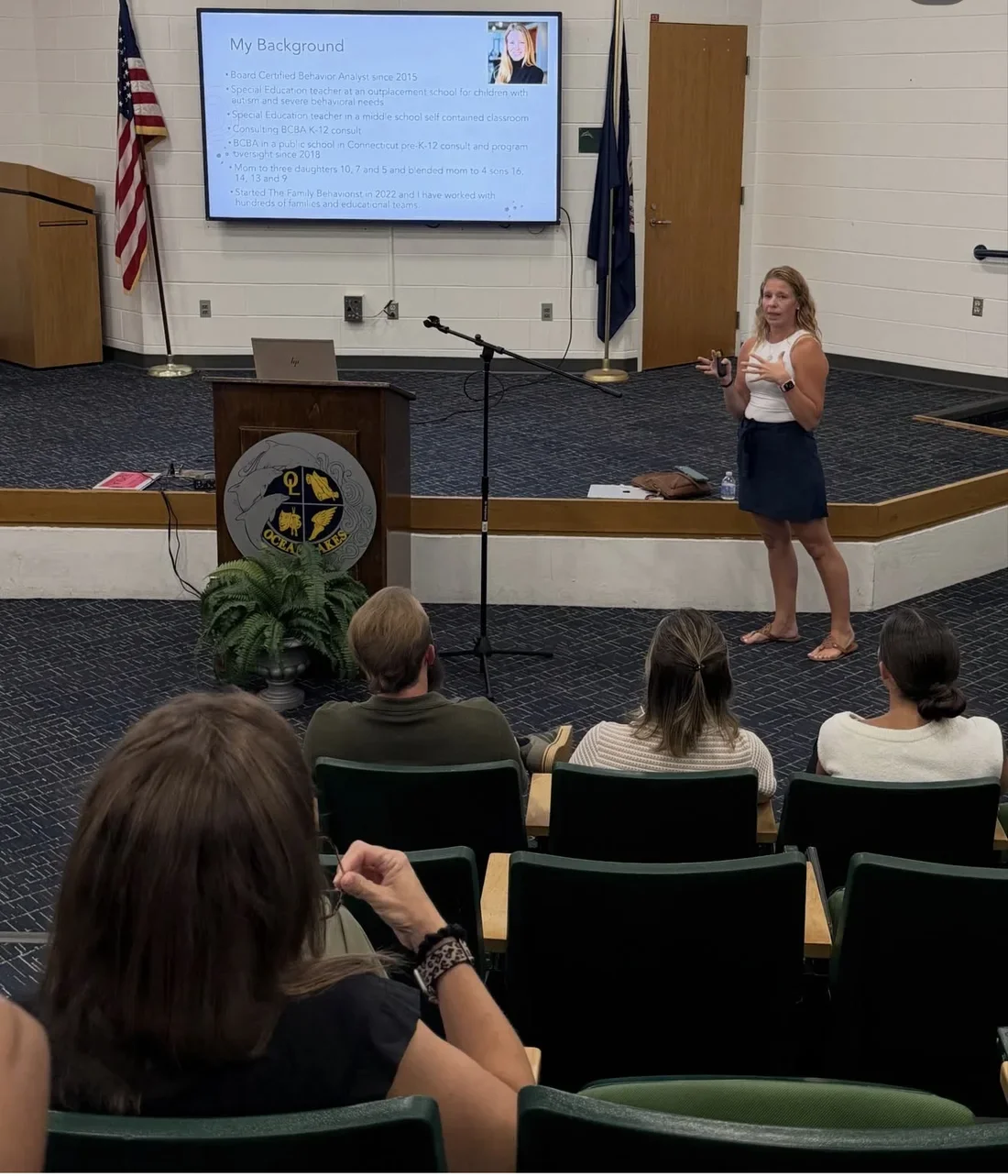 Mandy presenting to a school audience in an auditorium, with her background slide on screen