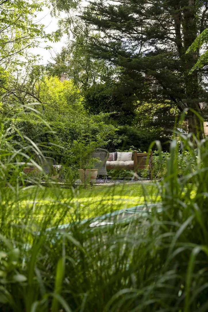 A backyard garden with lush green grass and dense trees. There is an outdoor seating area with a cushioned bench, a wicker chair, and potted plants, creating a cozy, shaded retreat.
