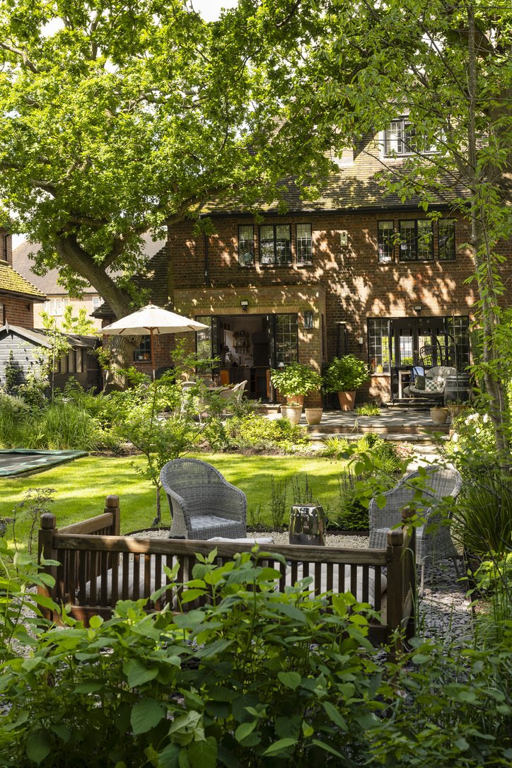 A lush green backyard with a large tree providing shade, a wooden house in the background, and outdoor furniture including chairs and a small table in the foreground.