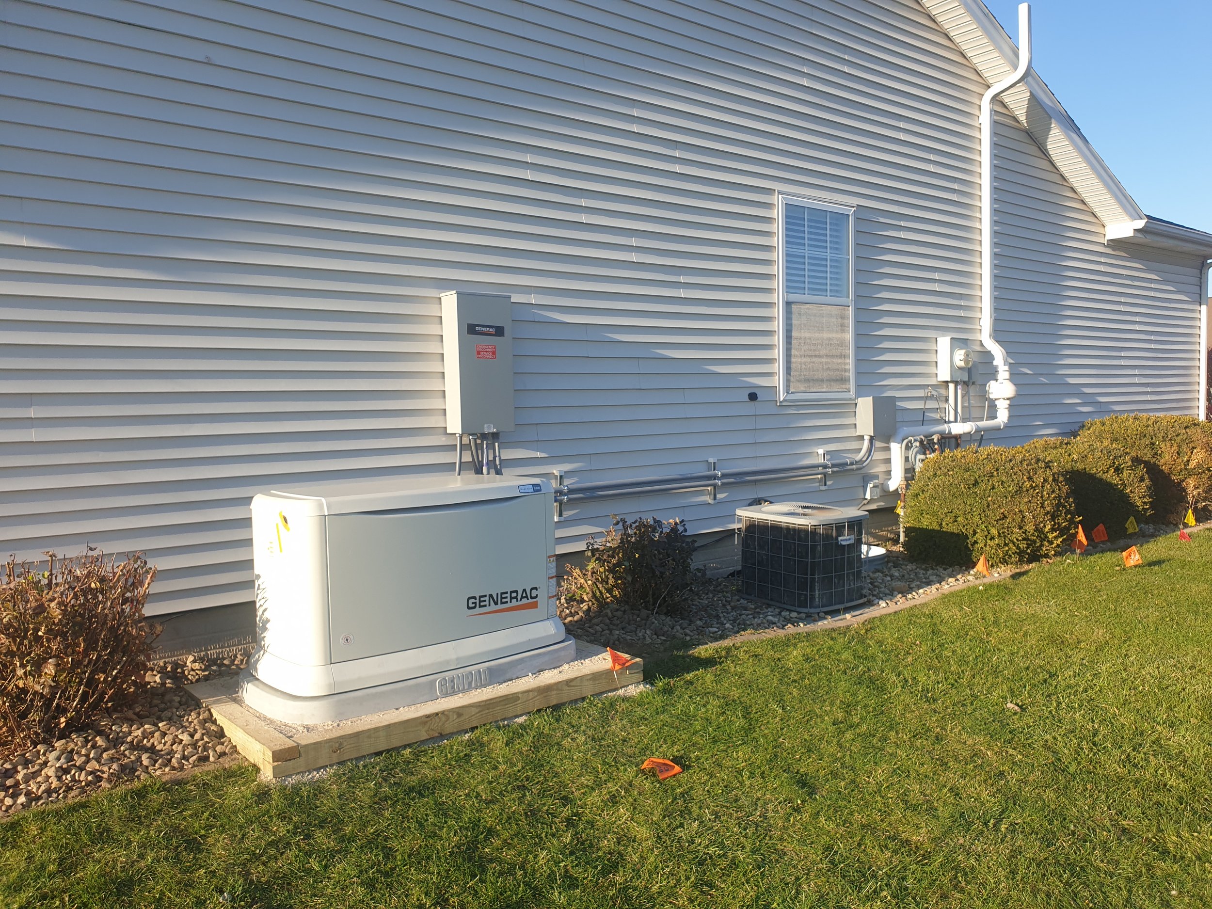 Side of a house with white vinyl siding, a window with white blinds, and a white gutter system. Several outdoor HVAC units and generator equipment are in the yard, surrounded by small rocks and bushes, with a green lawn and a clear blue sky in the background.