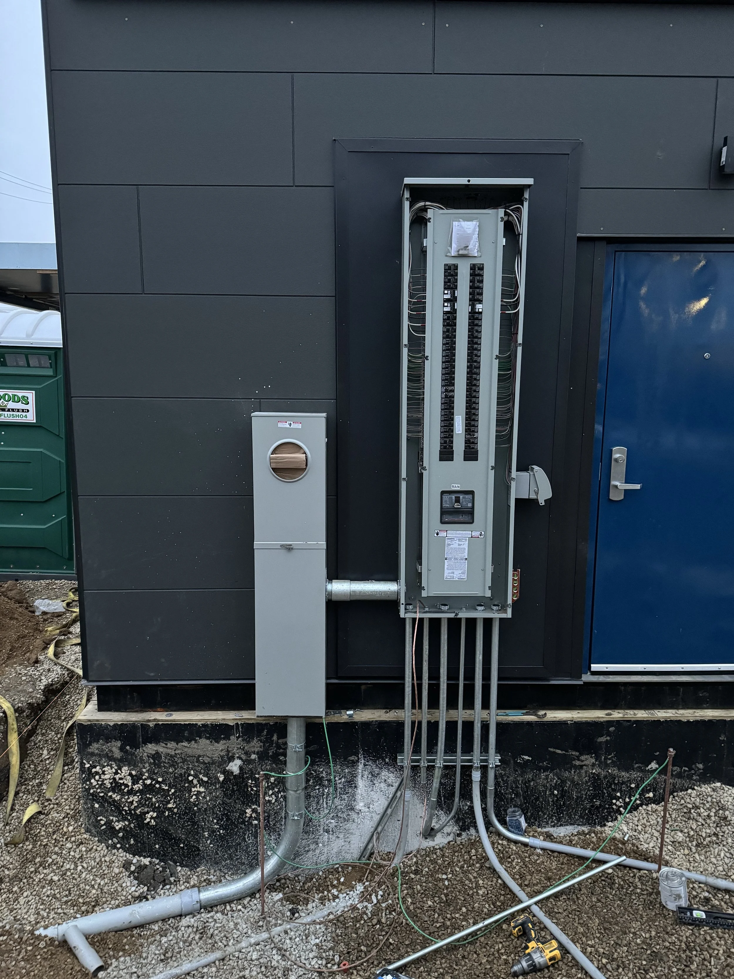 Electrical panel with wiring and pipes being installed outdoors on a construction site.