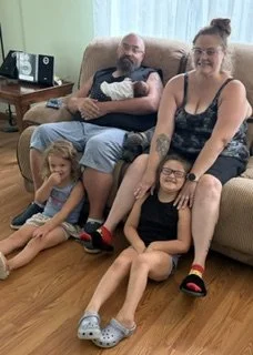 My granddaughter and her husband and their 3 girls our great granddaughters. A family sitting together in a living room; a man holding a newborn baby , a woman with glasses, and two girls sitting on the floor.
