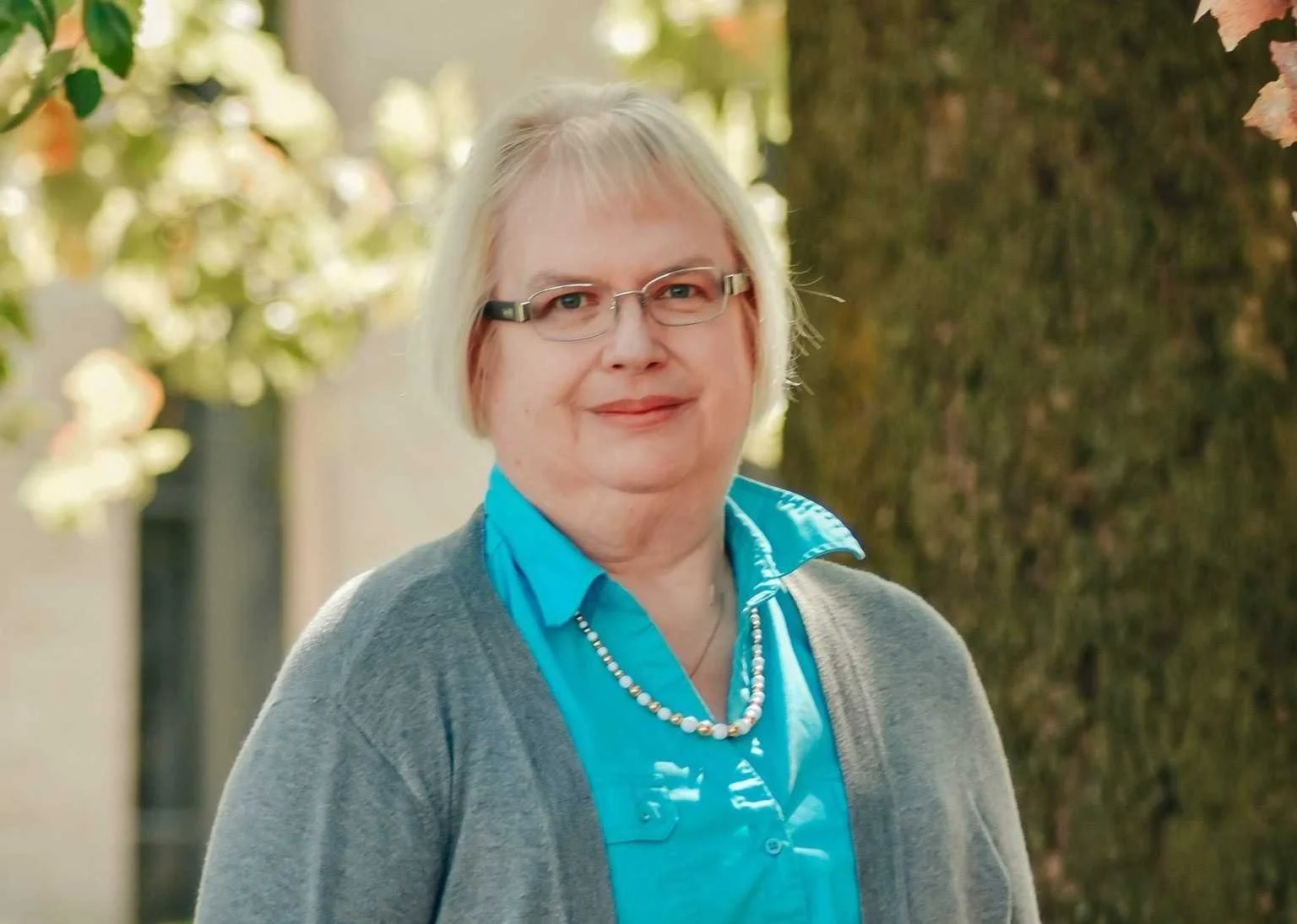 A middle-aged woman with light blonde hair, glasses, and fair skin stands outdoors near a tree with blurred green leaves in the background. She is wearing a blue button-up shirt, a gray cardigan, and a pearl necklace, smiling gently.