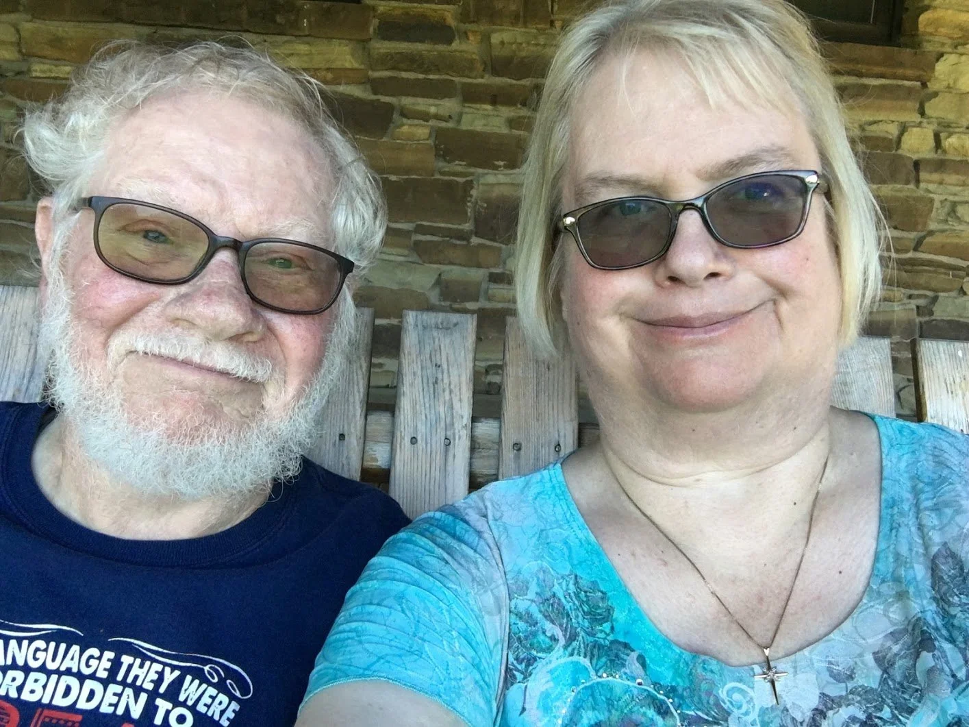 A smiling man with white hair and beard, wearing sunglasses and a navy blue t-shirt, taking a selfie with a smiling woman with blonde hair, wearing sunglasses and a blue-green patterned shirt, sitting on a wooden bench with a stone wall in the background.