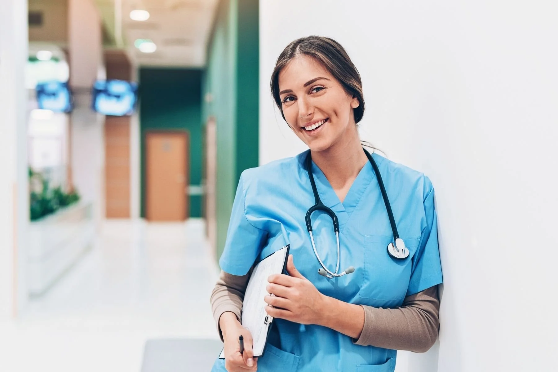 Smiling nurse in blue scrubs with stethoscope and clipboard in a hospital corridor.