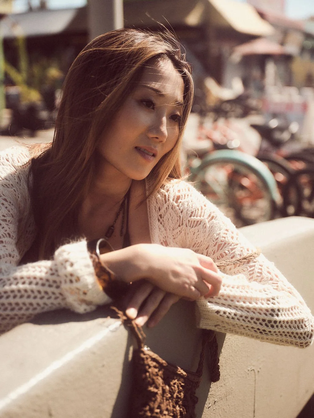 A woman with long hair sitting outdoors leaning on a ledge, smiling softly, with a background of bicycles and structures.