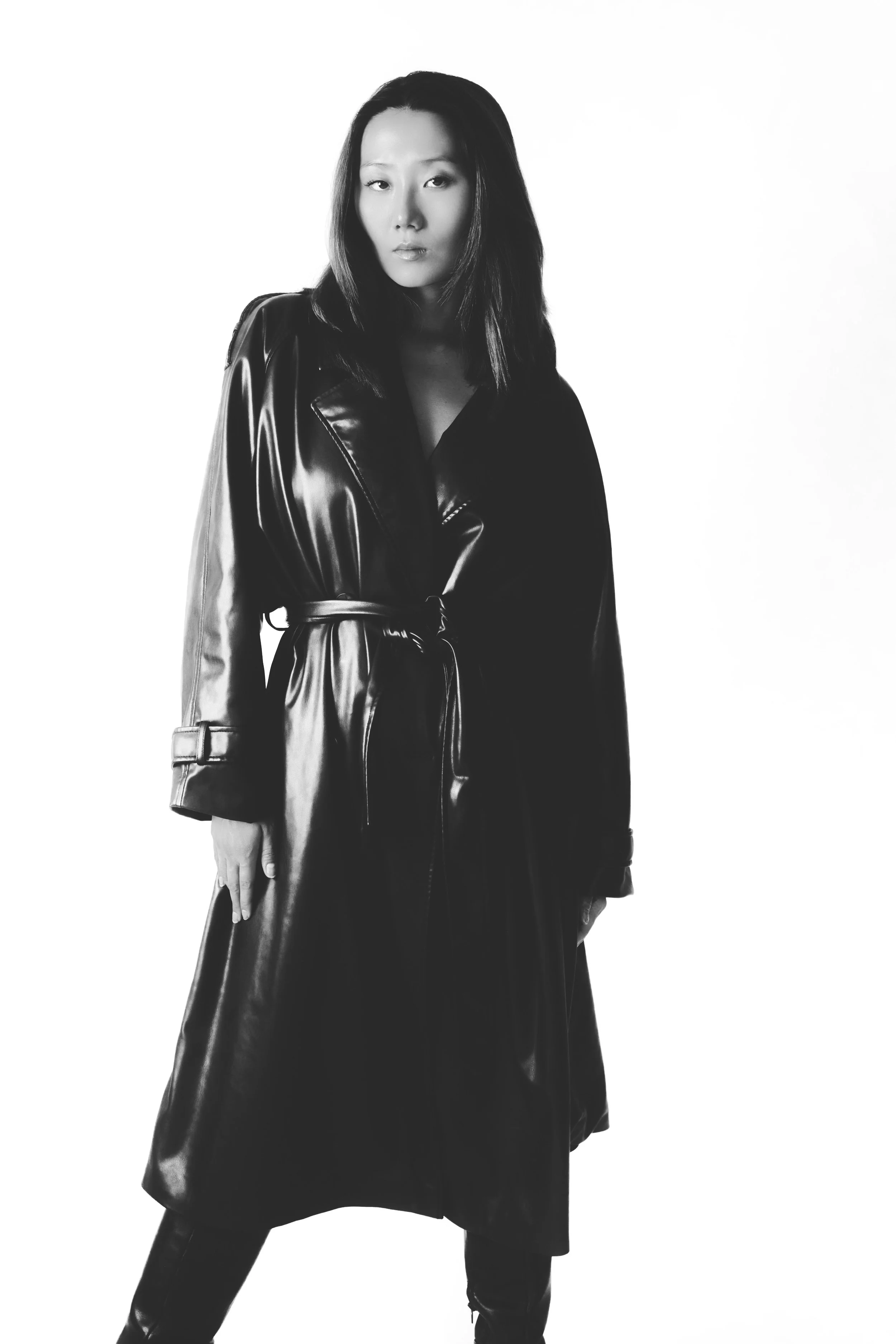 Black and white photograph of a woman wearing a leather trench coat, standing against a plain white background.