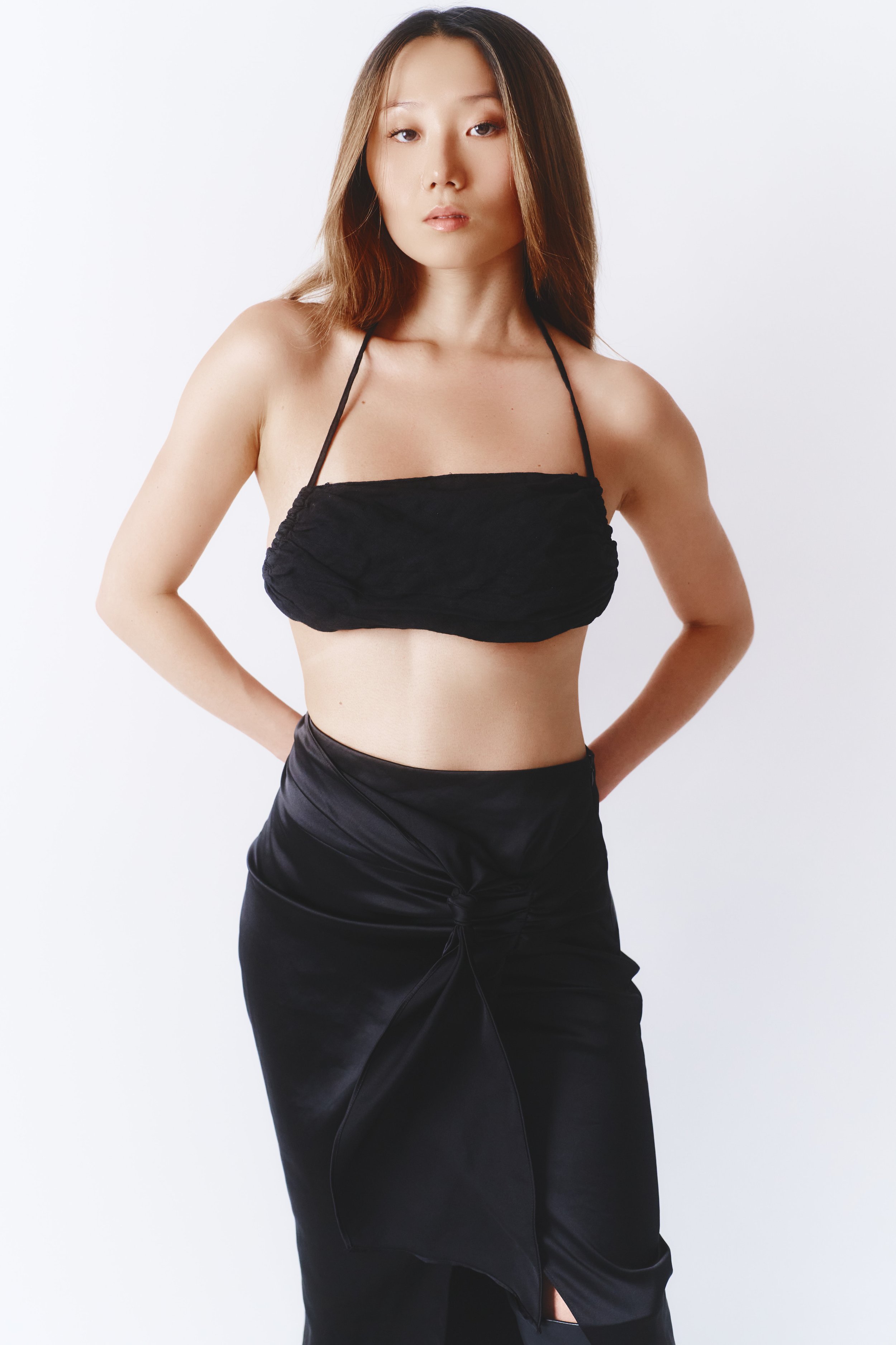 A woman with long, straight hair wearing a black strapless top and a black satin skirt stands against a plain white background.