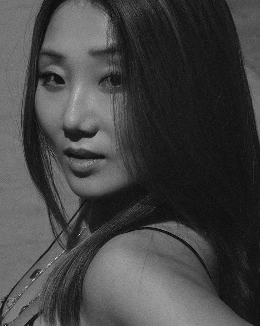 Close-up black and white photo of a woman with long straight hair, looking over her shoulder with a confident expression.