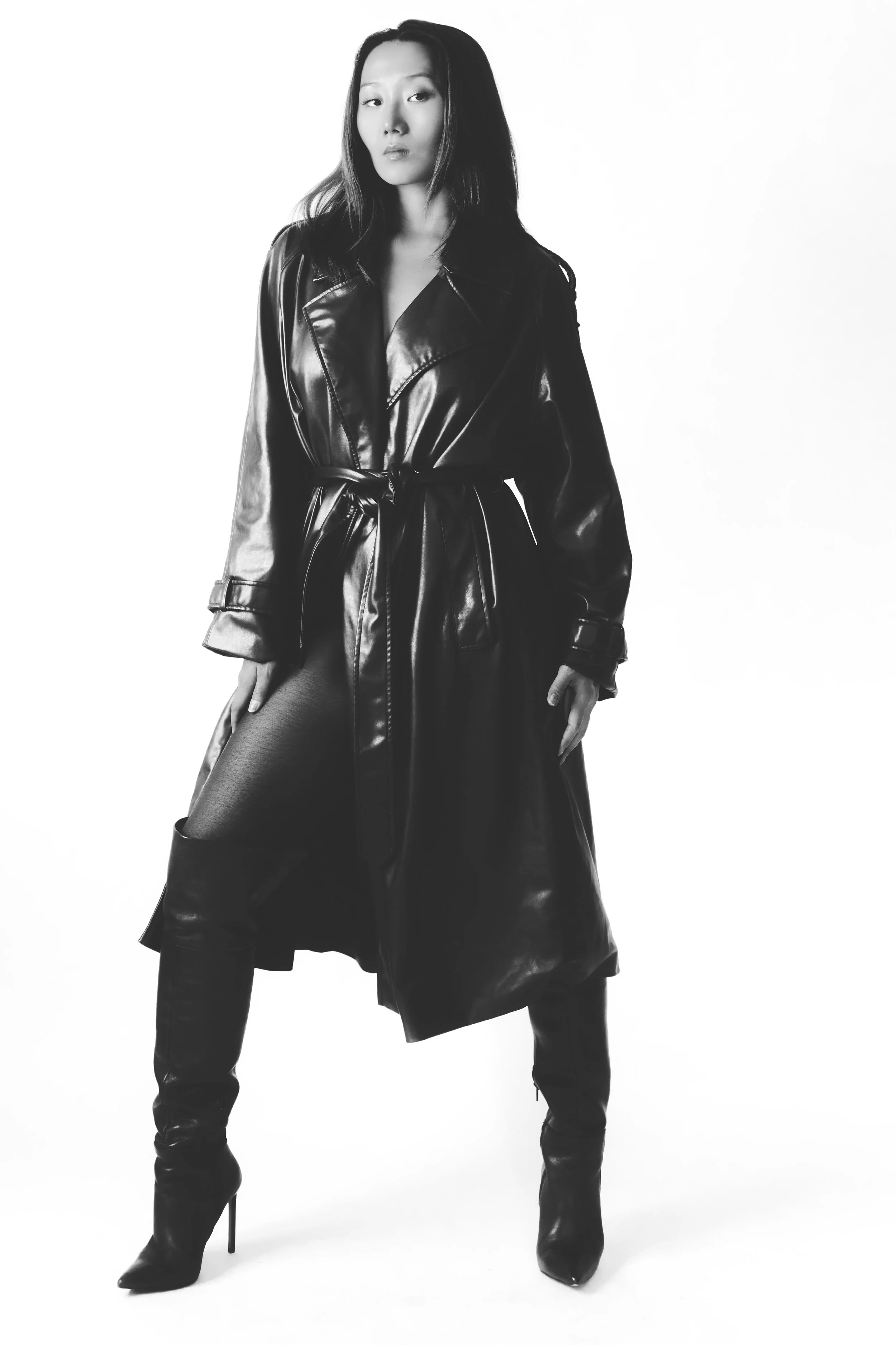 A woman in a long, shiny black leather coat, thigh-high black boots, posing against a white background.