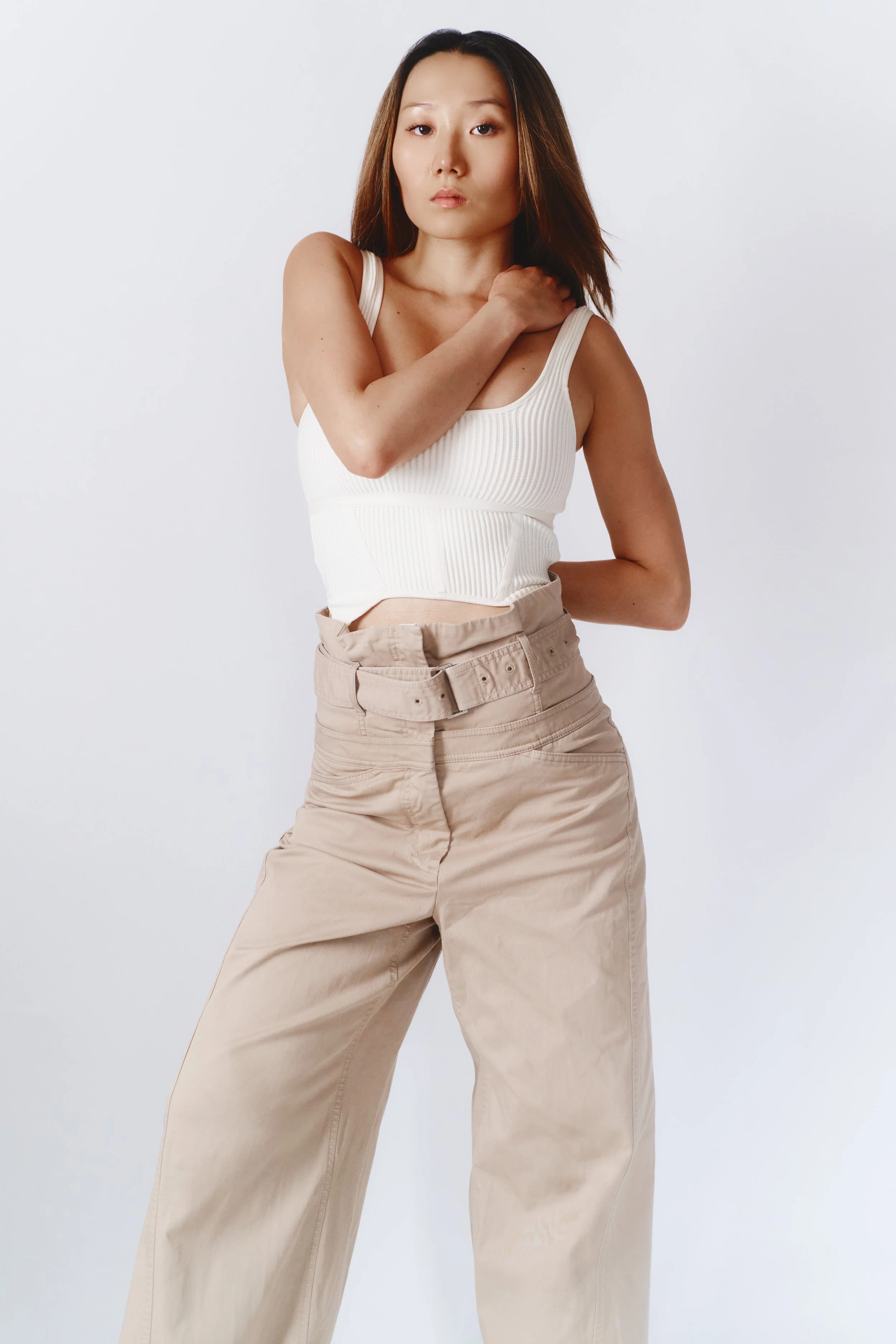 A woman with shoulder-length brown hair wearing a white ribbed tank top and beige wide-leg pants with a matching belt, posing against a plain white background.