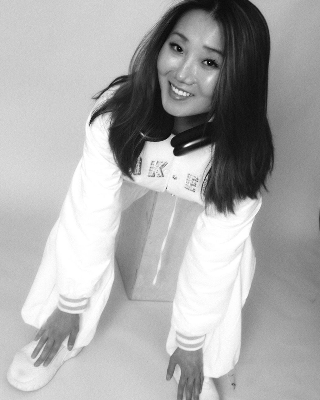 A young woman with long dark hair smiling, wearing a white hoodie with text, crouching with hands on knees, and a pair of headphones around her neck, against a plain background.