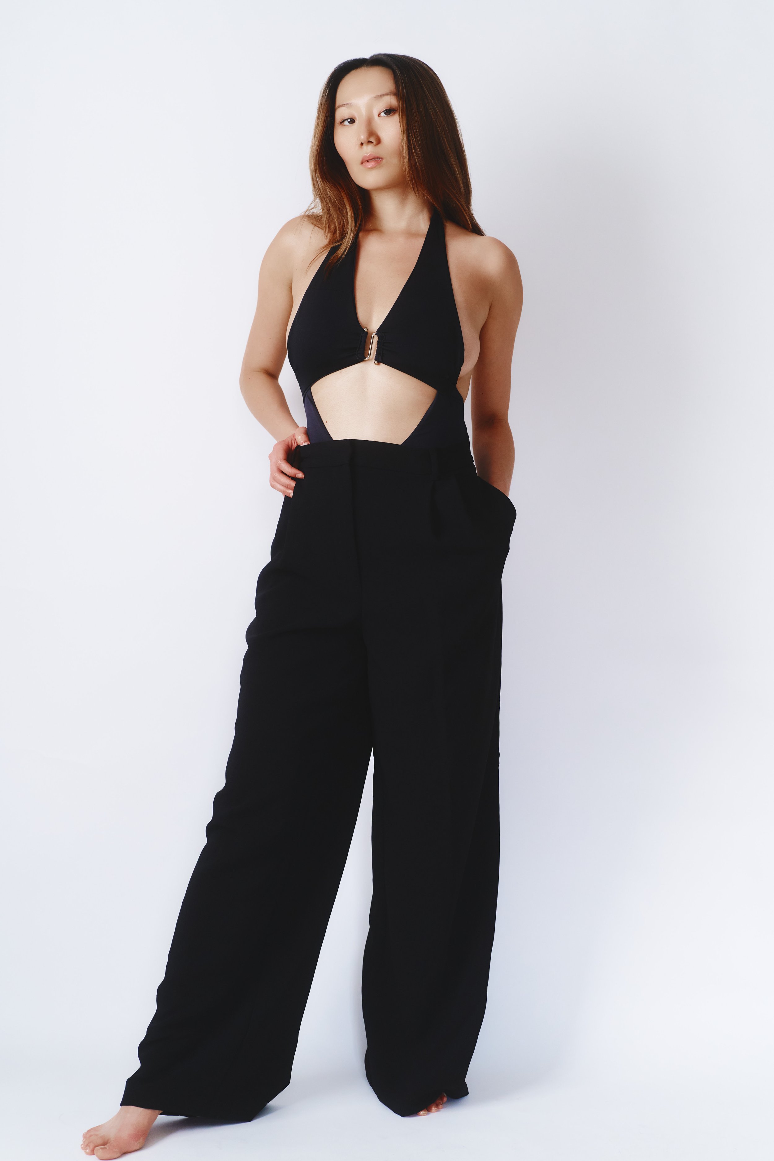 A woman with long hair wearing a black halter top with a zipper and large cutout, paired with black high-waisted wide-leg pants, standing against a plain white background.