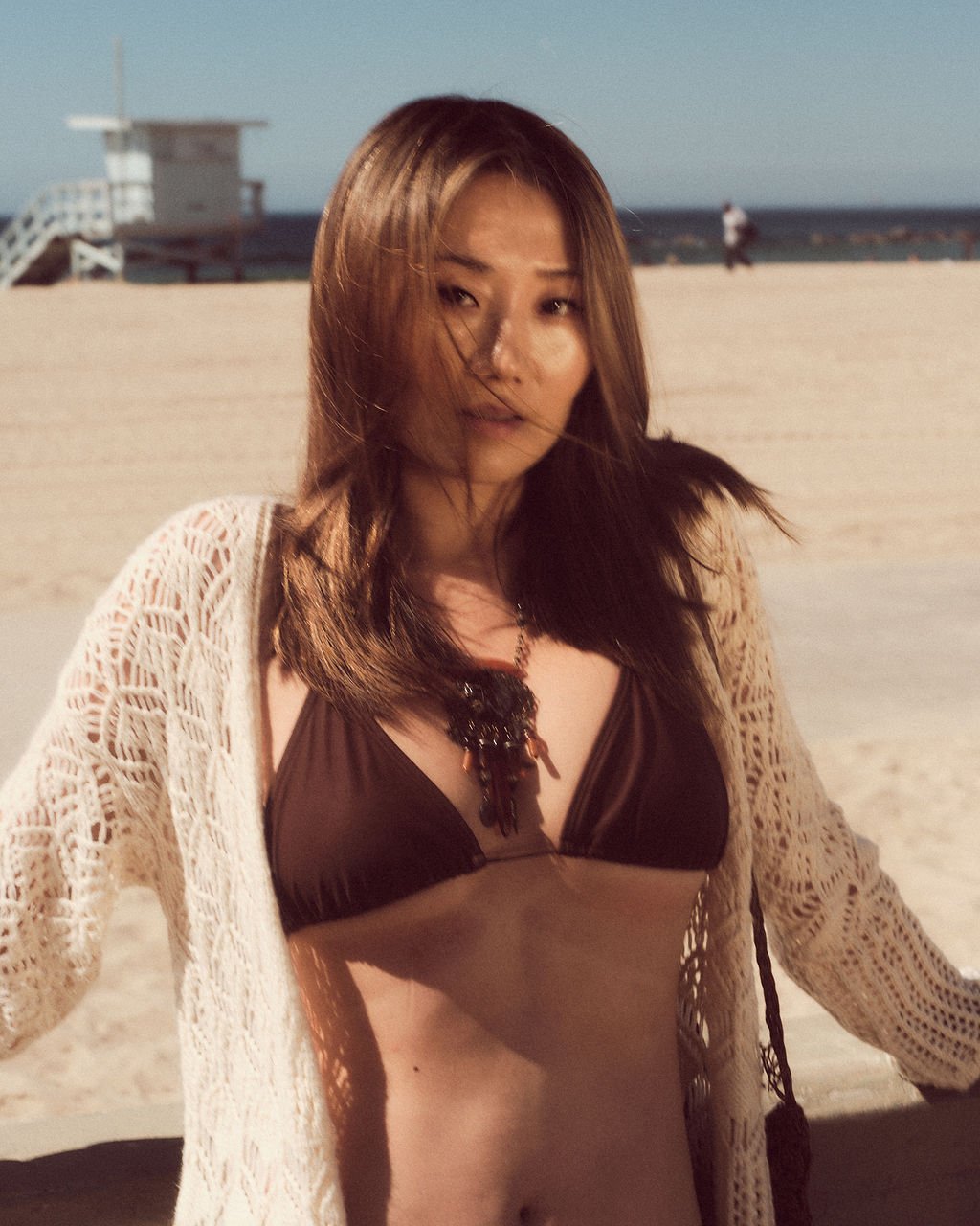 A woman in a bikini and a lace cover-up on the beach, with a lifeguard tower and ocean in the background.