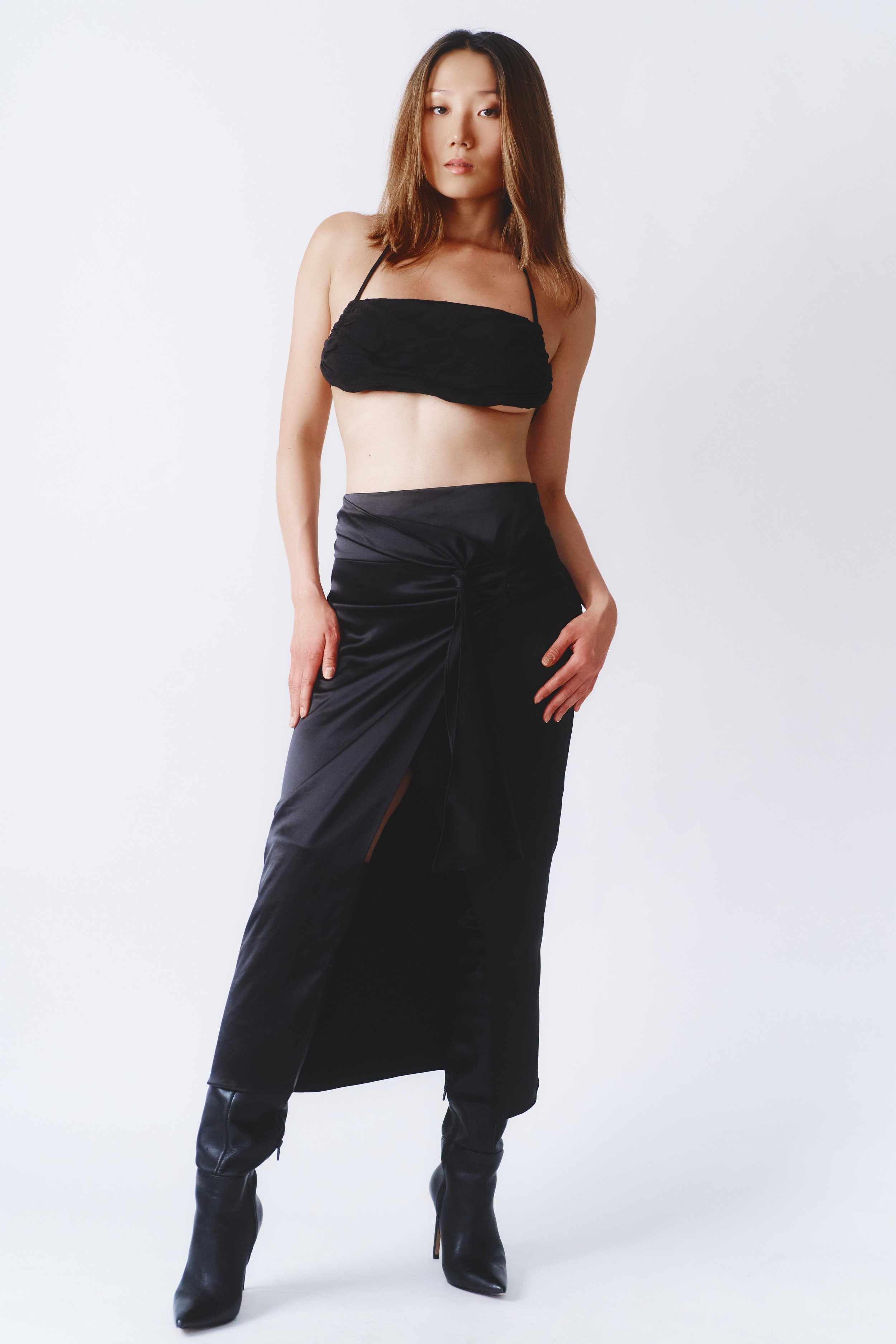 A woman wearing a black crop top, black skirt with a slit, and black high-heeled boots posing against a plain white background.
