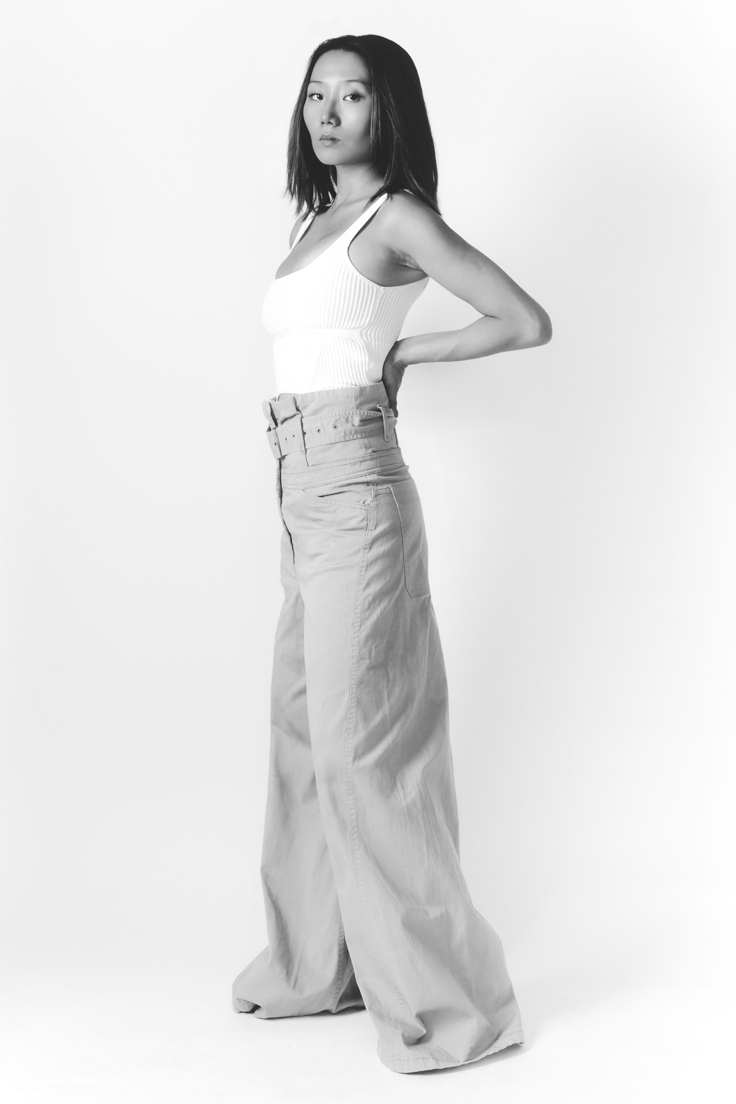 Black and white photo of a woman with shoulder-length hair, wearing a sleeveless top and wide-leg pants, standing against a plain background.