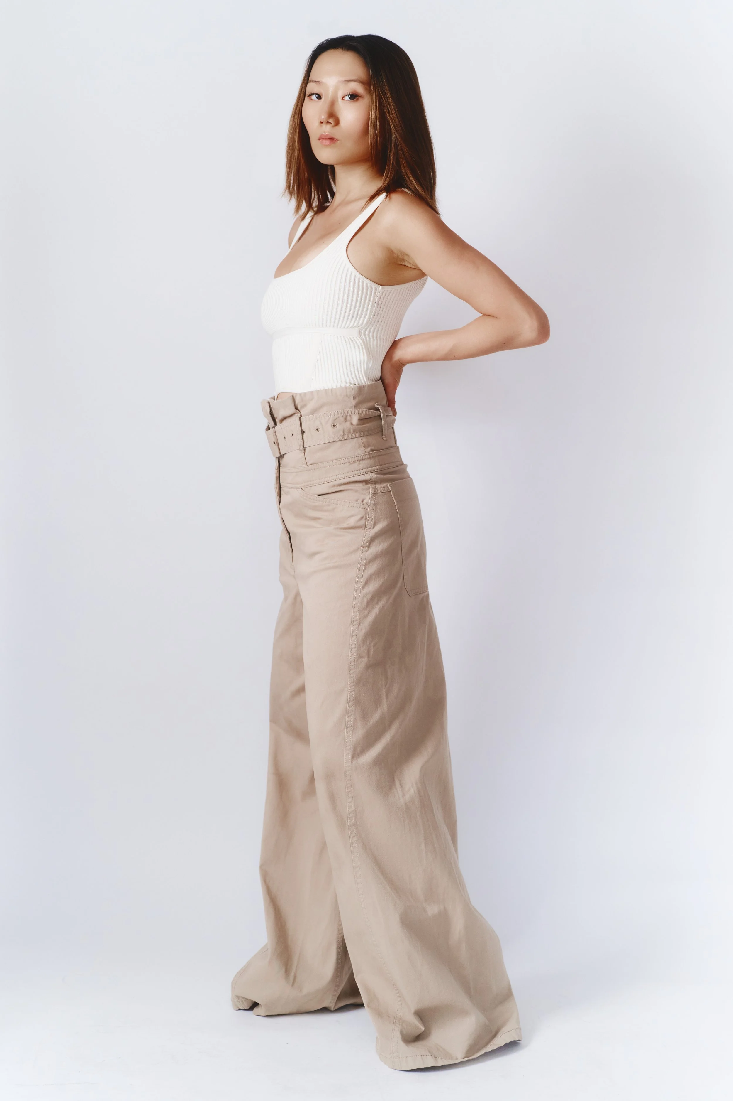 A young woman with shoulder length dark hair, wearing a white ribbed tank top and wide beige pants, standing against a plain white background, posing with her left hand on her hip and looking at the camera.