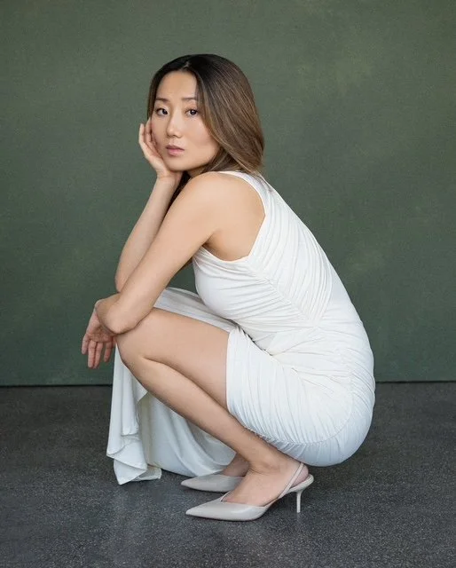 A woman in a white dress squatting with her right hand on her cheek, looking at the camera, against a plain green background.