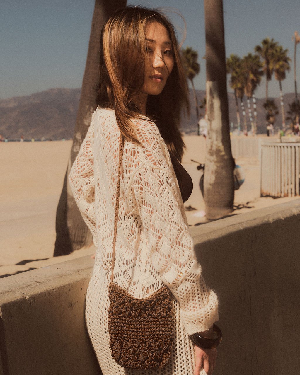 A woman standing outdoors on a beachside walkway with palm trees, mountains, and people in the background. She has shoulder-length brown hair, wears a white lace crochet cardigan, and carries a brown woven bag.