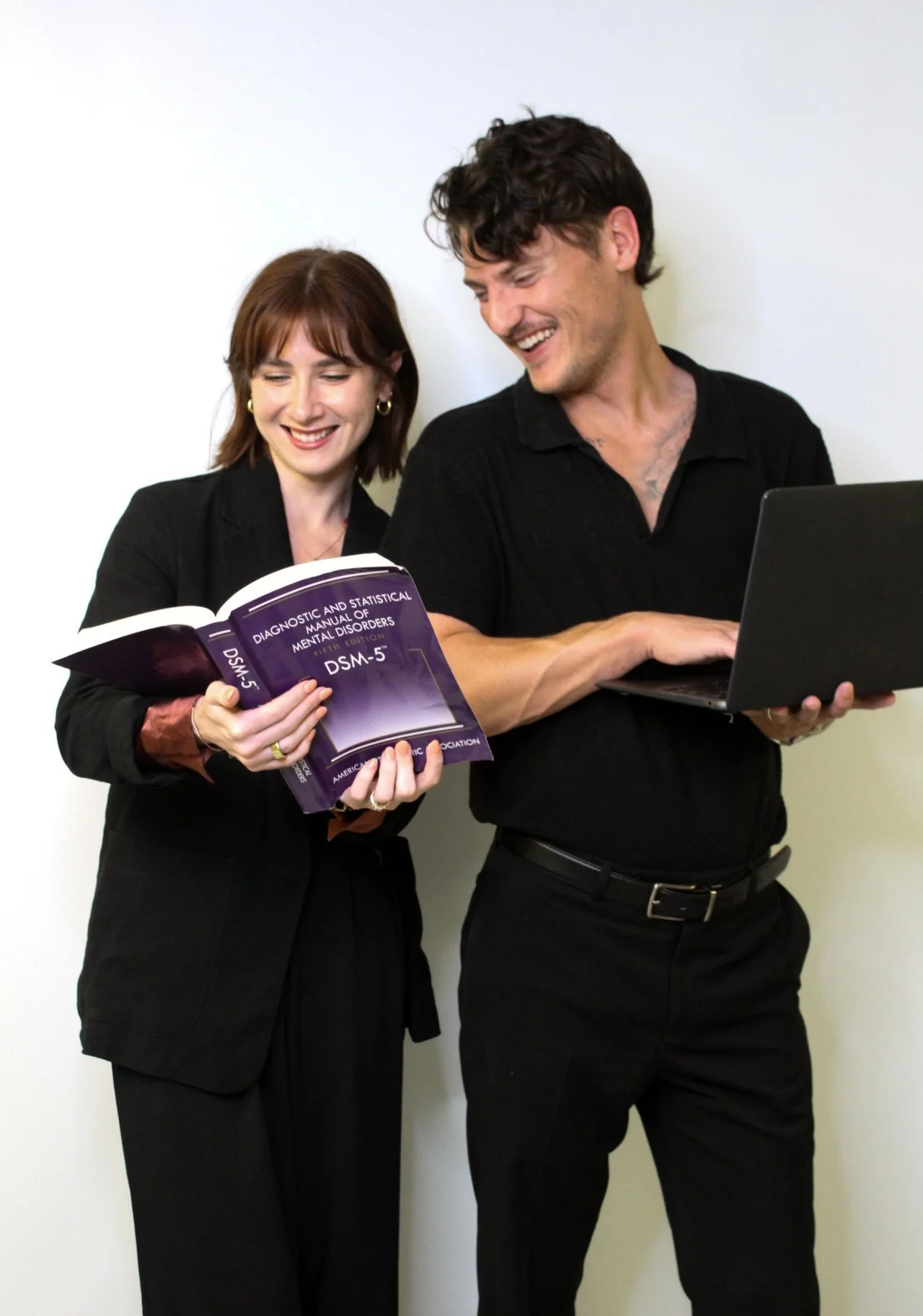 A woman and a man standing together, smiling, looking at a book and a laptop. The woman holds a book titled 'Diagnosing and Statistical Manual of Mental Disorders'.