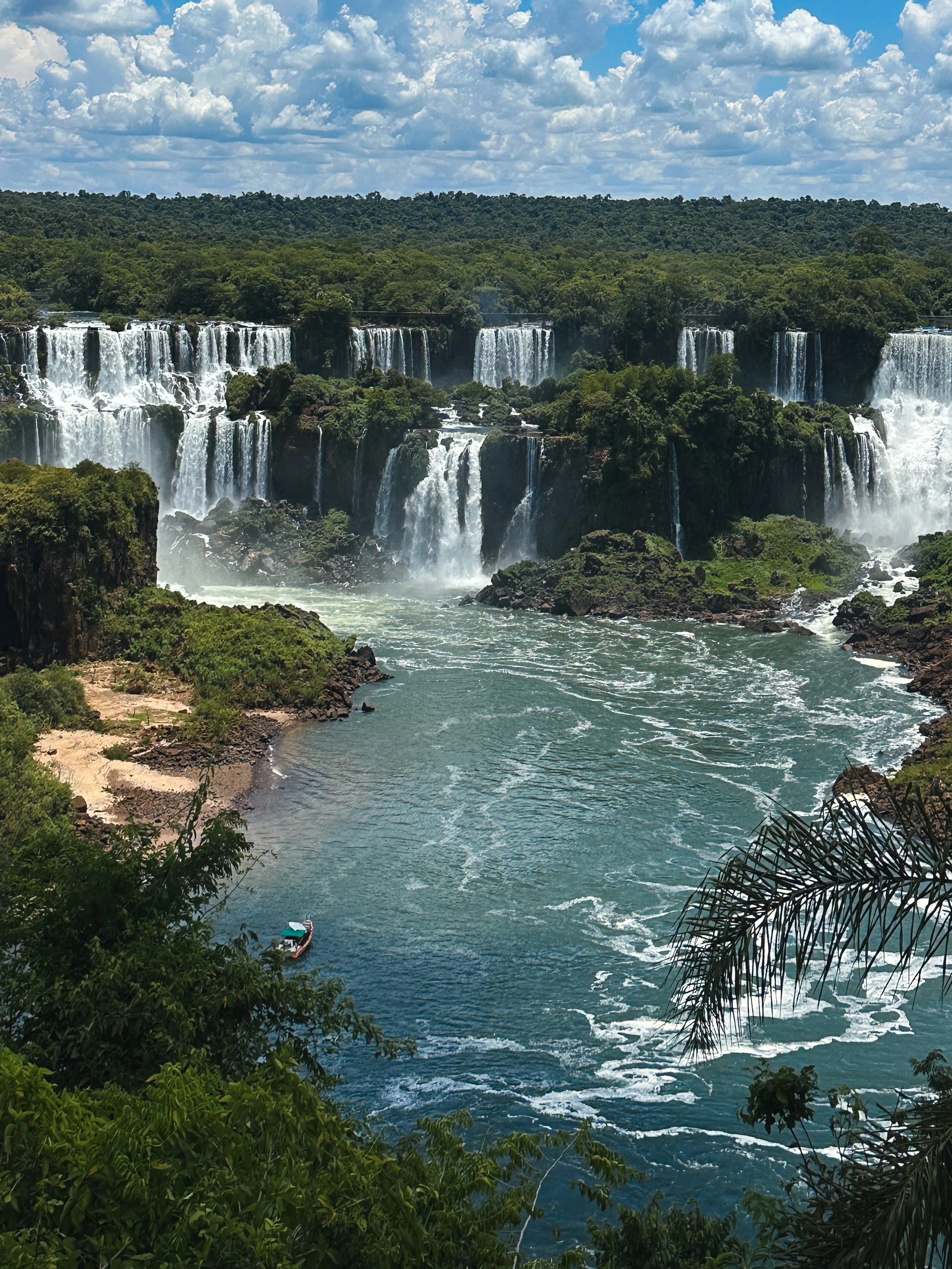 A scenic view of waterfalls in a lush green jungle, with a boat on the river and a partly cloudy sky overhead.