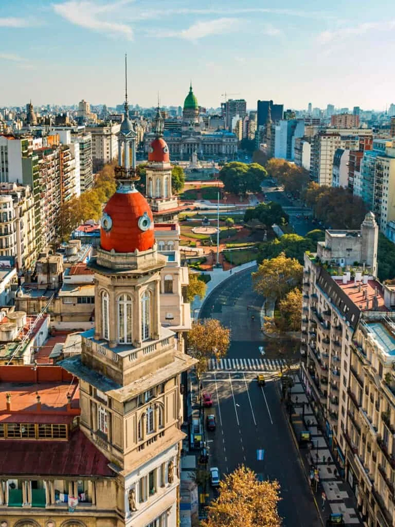 Bird's eye view of a cityscape with historic and modern buildings, green parks, and streets, with a prominent clock tower with a red roof in the foreground and a domed government building in the background under a clear sky.