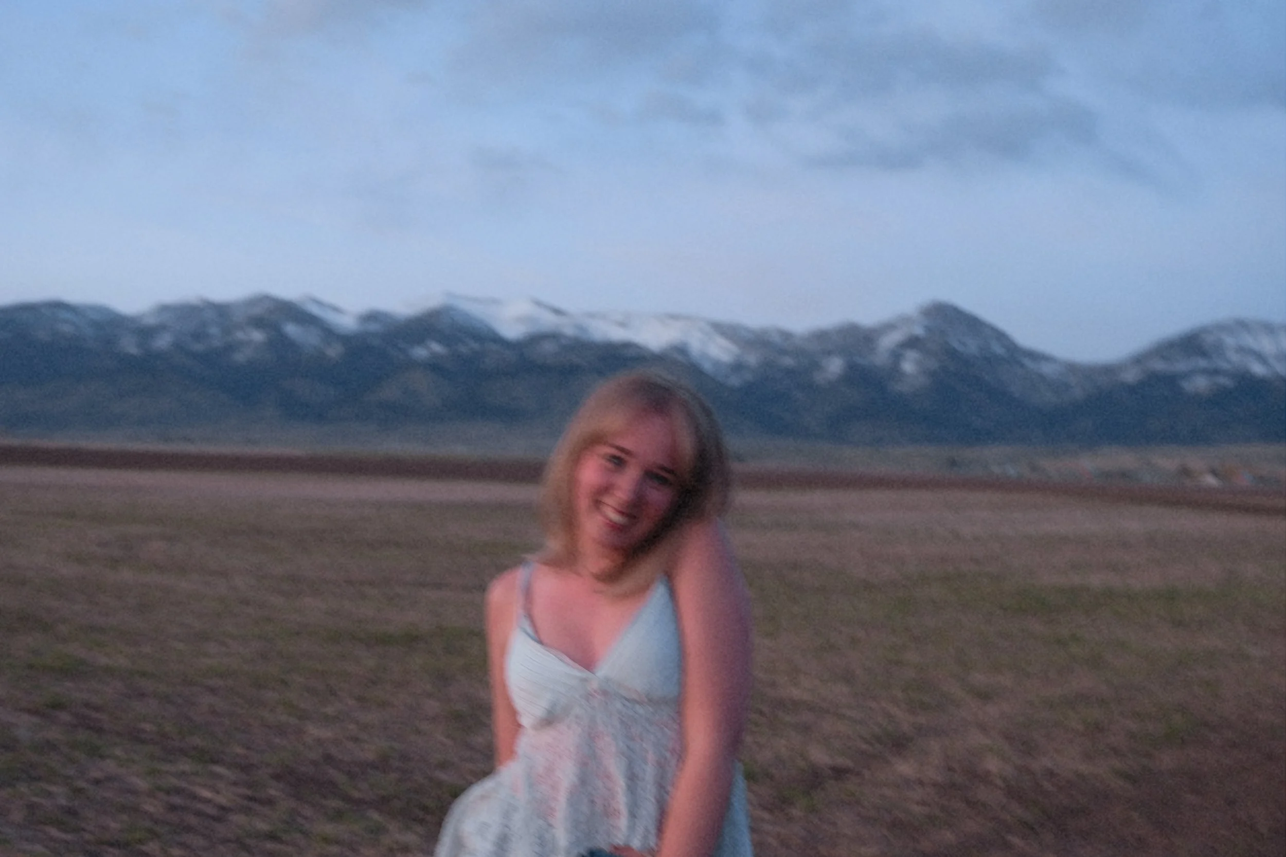 A smiling young woman with blonde hair in a sundress standing in an open field with mountains in the background during dusk.