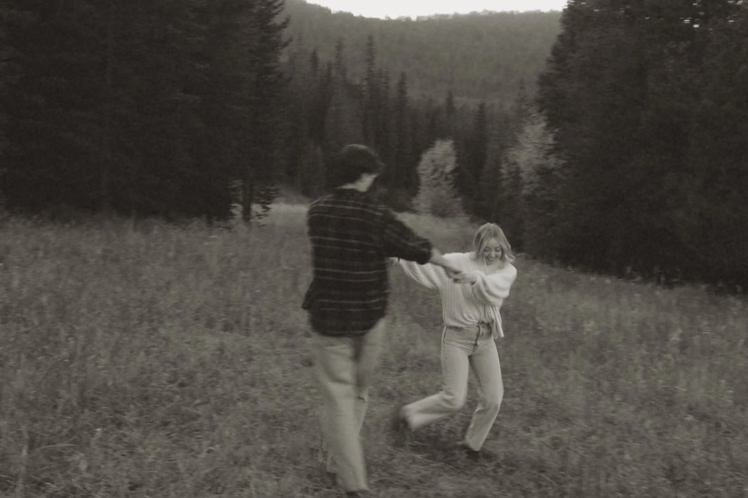 A black and white photo of a young man and woman playing in a grassy field with trees and mountains in the background. The woman is smiling and holding onto the man's arm.