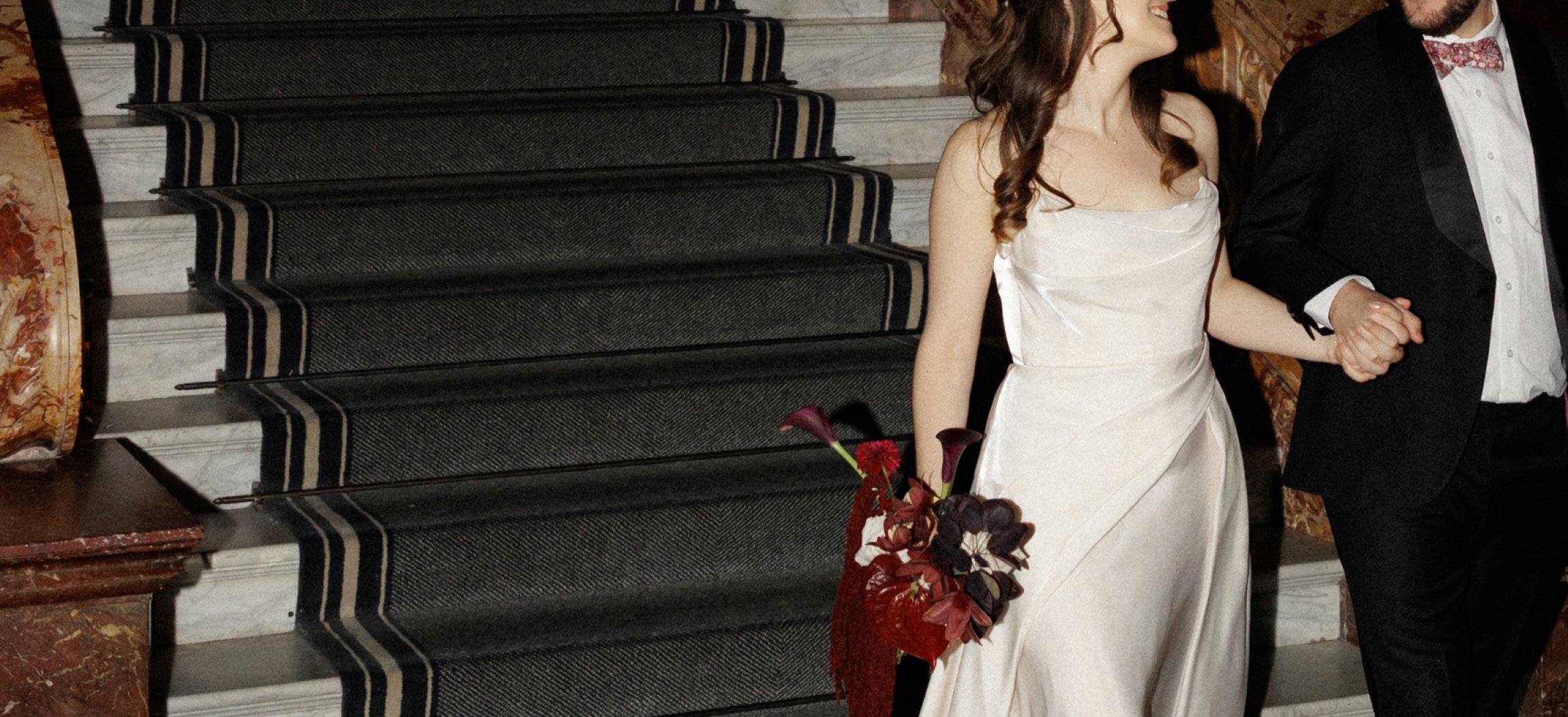 Bride and groom walking down marble stairs, the bride holding a modern bouquet in deep burgundy tones with calla lilies, anthuriums, dahlias and roses.