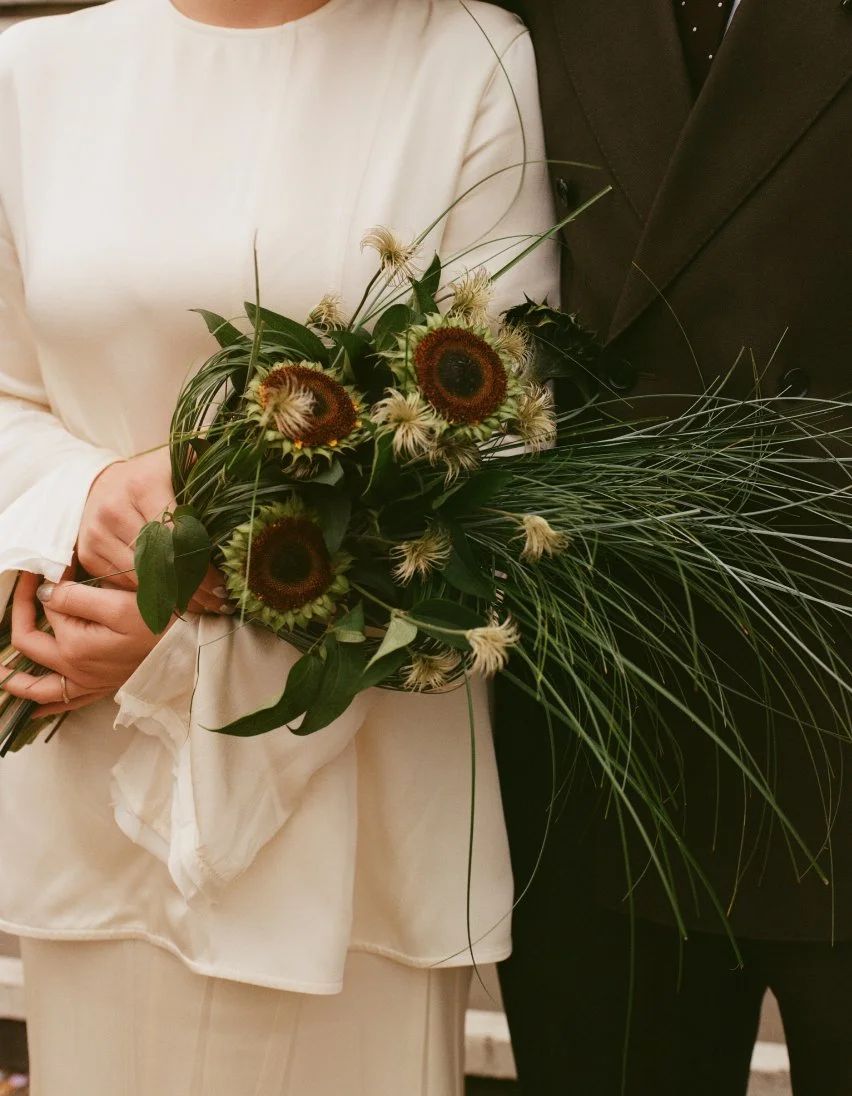 Close-up of a bride holding a modern bouquet with sculptural grasses, sunflower seed heads and clematis.