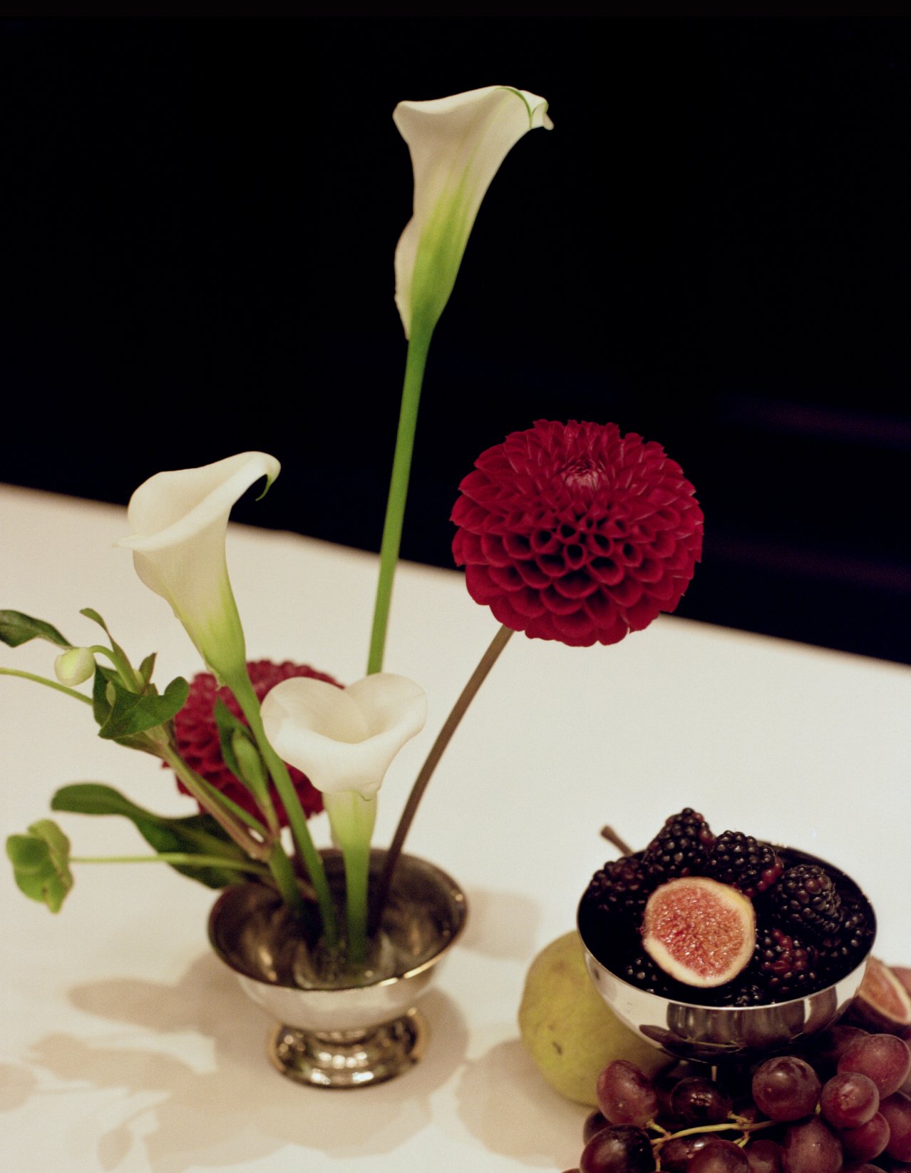 Ikebana-style arrangement in silver bowls with white calla lilies, dark red dahlias and hellebore, next to a small silver bowl of fruit.