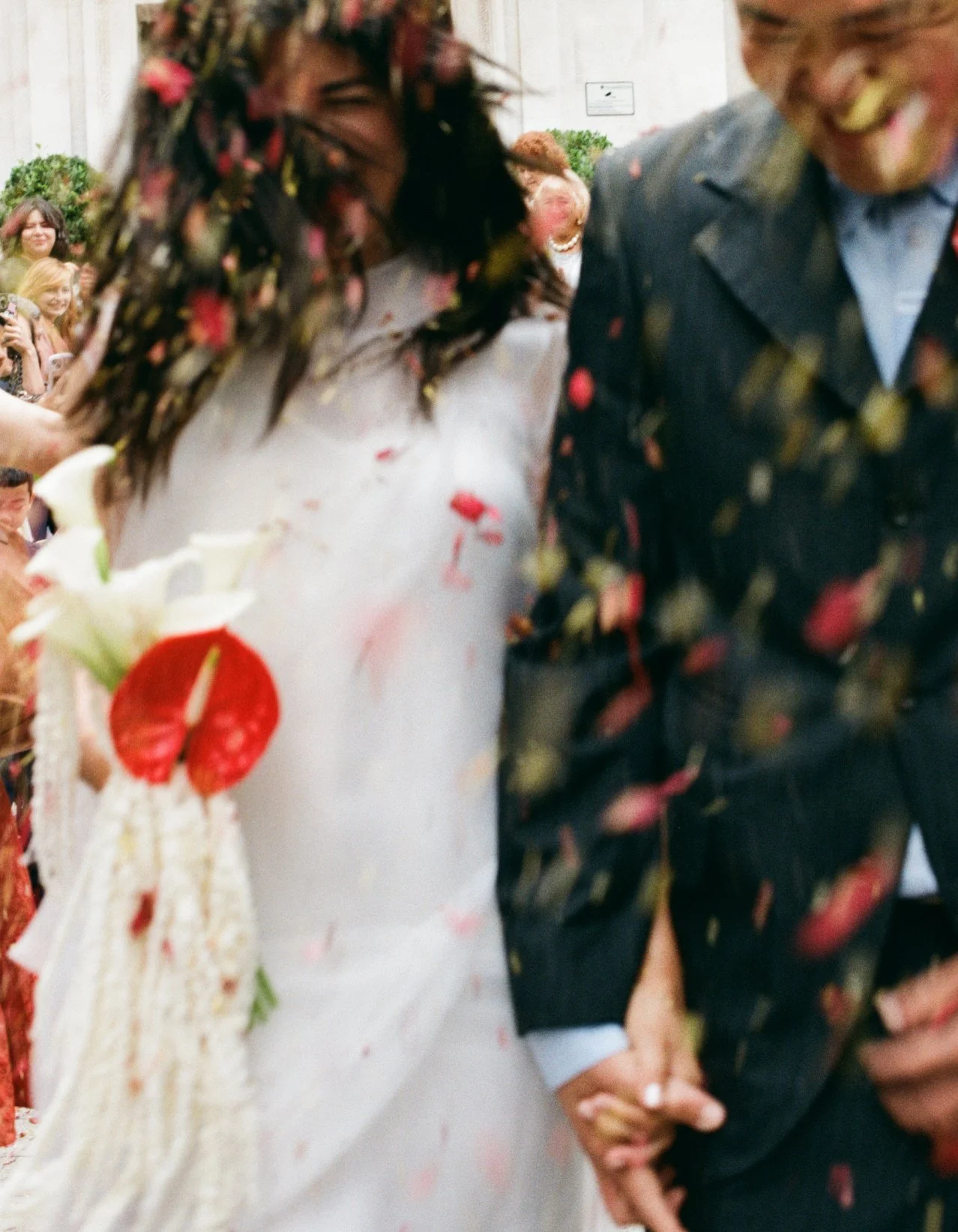 Couple walking through confetti after their wedding ceremony, the bride holding a bouquet with white calla lilies and red anthurium.