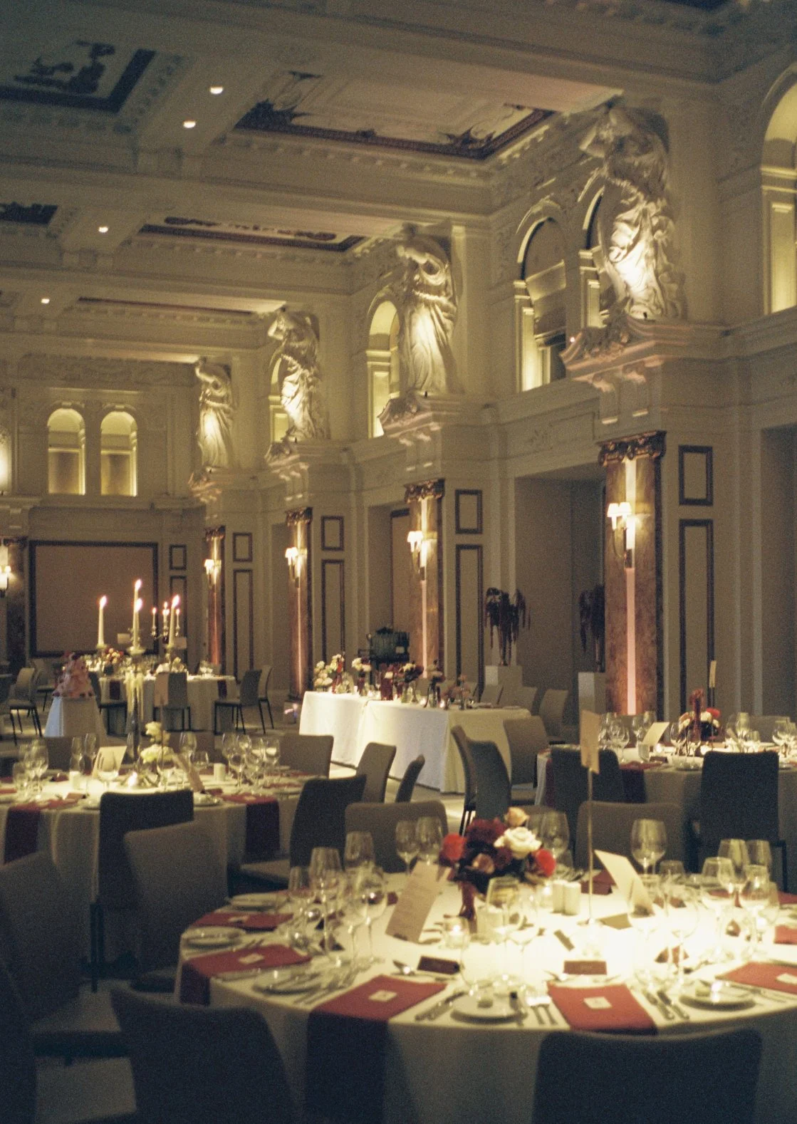 Wedding reception with round tables, candles and burgundy floral centrepieces in an ornate ballroom.
