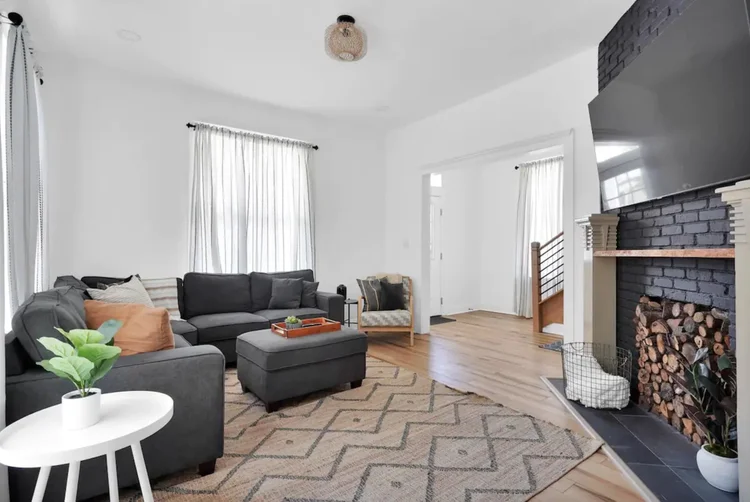 Living room with a gray sectional, a painted brick fireplace with stacked firewood, and a wall-mounted TV