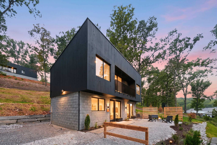 Two-story cabin with black vertical siding over a gray lower level, lit windows, and a balcony, beside a gravel patio with chairs around a fire pit at dusk