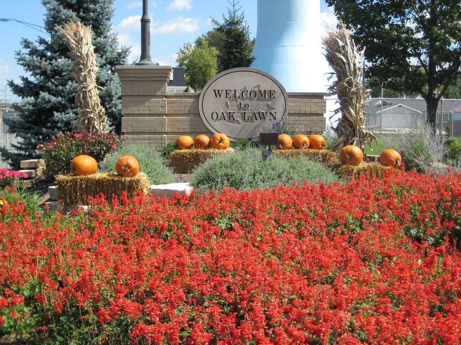 Welcome to Oak Lawn sign on a brick base with pumpkins and hay bales, red salvia in front, and a water tower behind