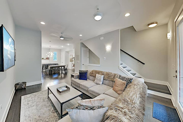 Modern living room with an L-shaped sofa, a coffee table, and a wall-mounted TV, leading to a dining area and staircase
