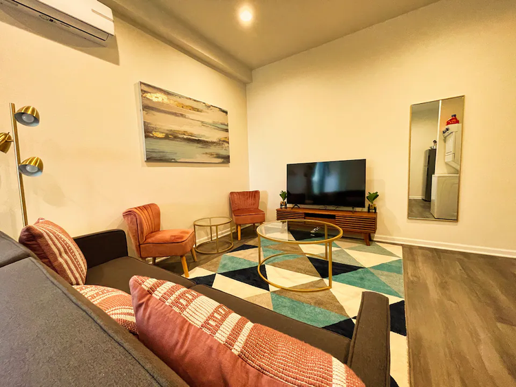 Living room with a sofa and patterned pillows, two orange accent chairs, a geometric rug, and a TV on a wood console