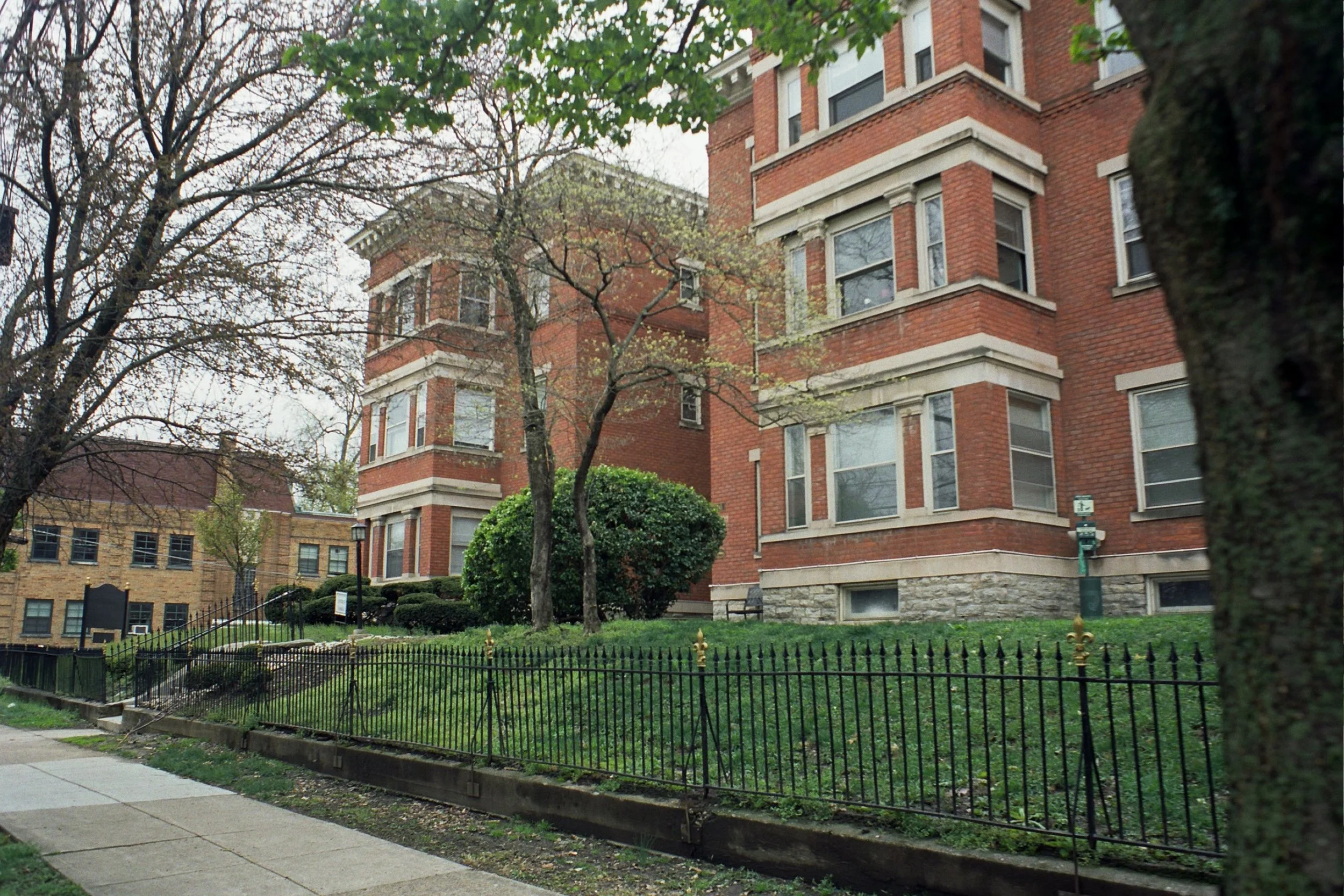 Red brick vintage apartment building behind an iron fence and a green lawn