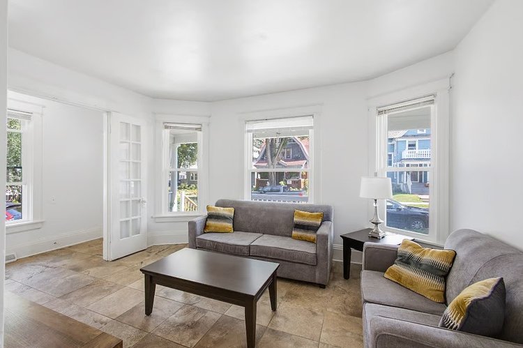 Living room with two gray sofas, yellow accent pillows, a dark wood coffee table, and tall windows facing the street
