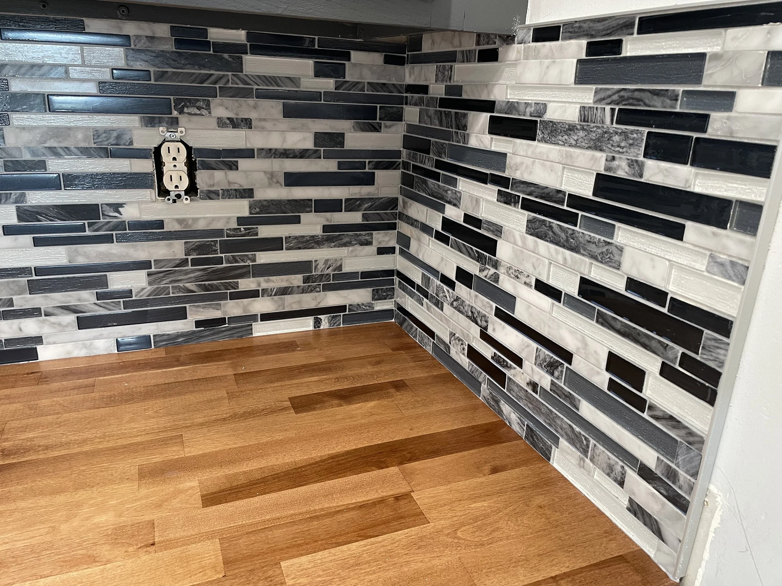 Close-up image of a bathroom wall with mosaic tile pattern in black, white, and gray marble, and a double electrical outlet with no cover plate, located near a wooden floor.