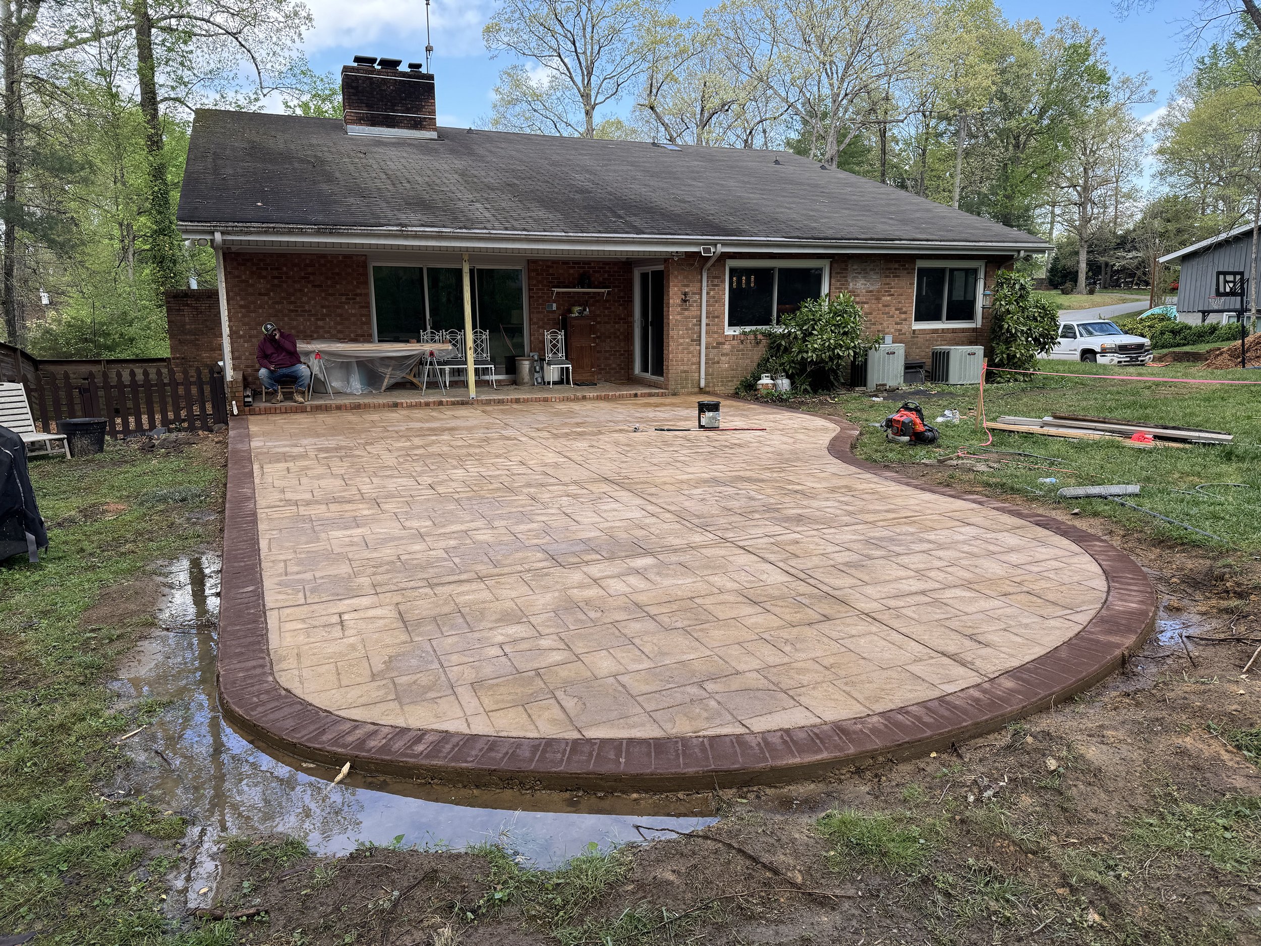 Backyard with newly paved brick patio, shrubbery, and a brick house with large windows and a sliding door. Worker is kneeling on the patio, working on finishing touches; tools and construction materials are scattered around.