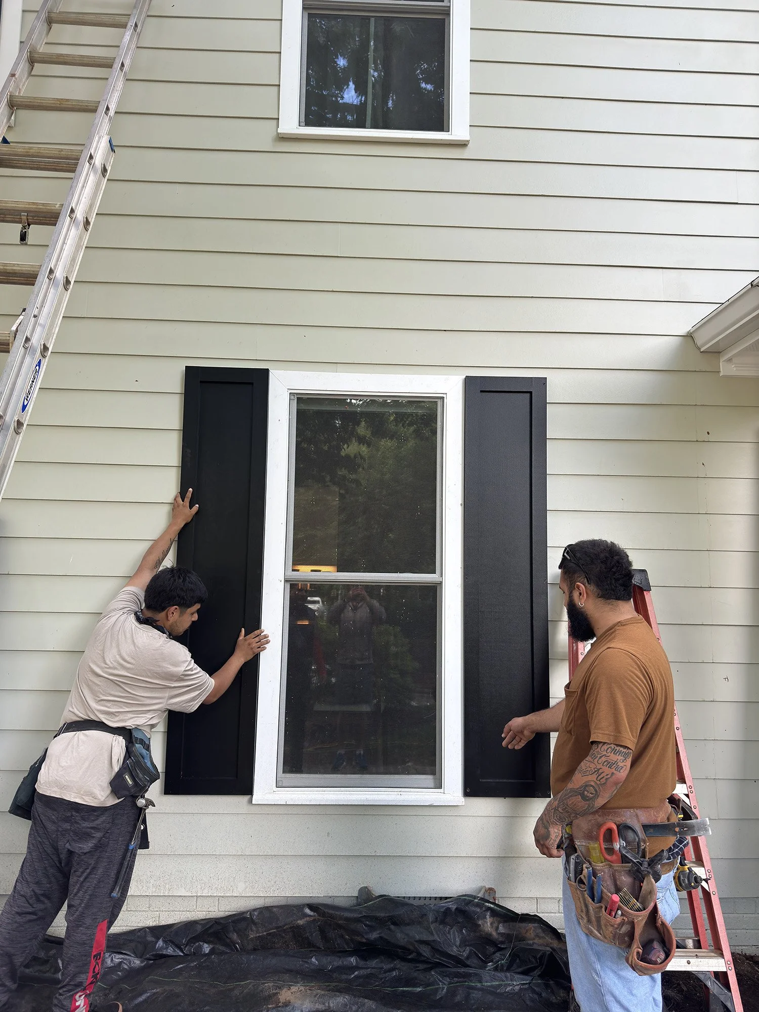 Three men installing or repairing a window on the exterior of a house. One man is on the left, leaning in and touching the window frame, another standing on the right with a tool belt and a tool in his hand, and a third man is partially visible in the mirror reflection. A ladder is leaning against the house, and a black tarp is on the ground underneath.