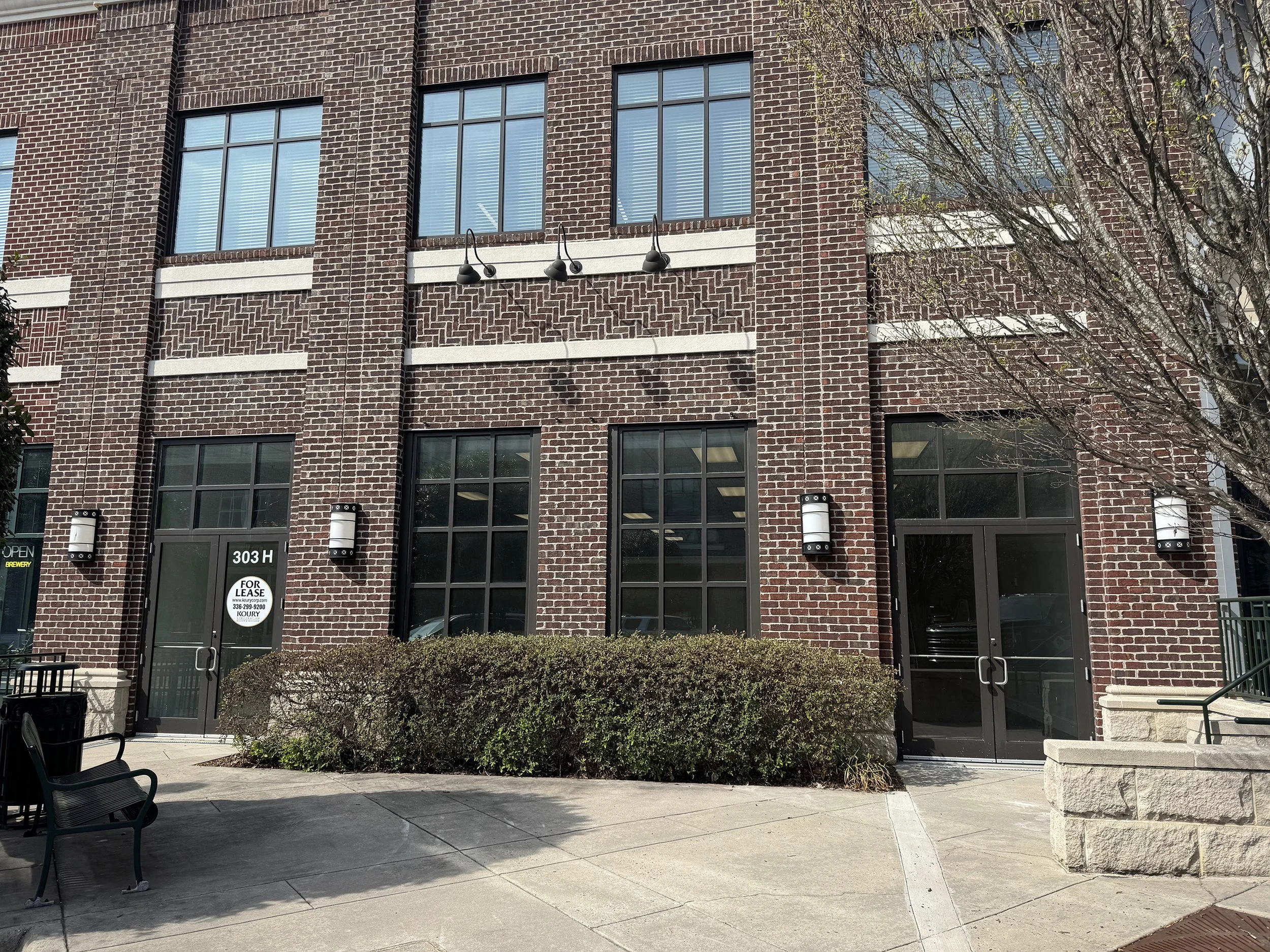 A multi-story brick building with several large windows, mounted exterior lights, and a small bush in front. There is a sign on the door that says 'For Lease' with contact information.