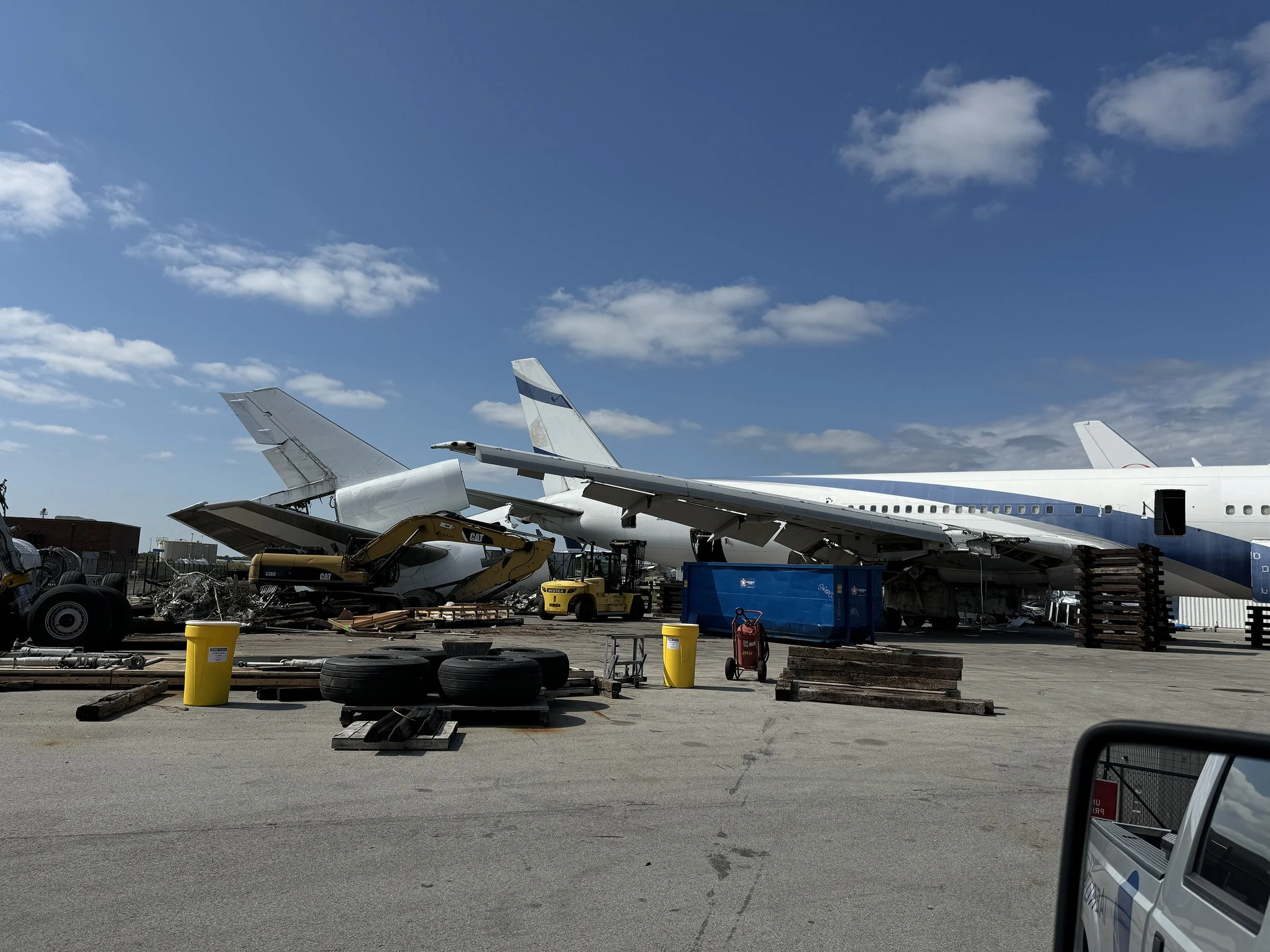 Aircraft parts and wreckage stored outdoors under a partly cloudy sky, with yellow trash bins, tires, wooden pallets, and construction equipment in the foreground.