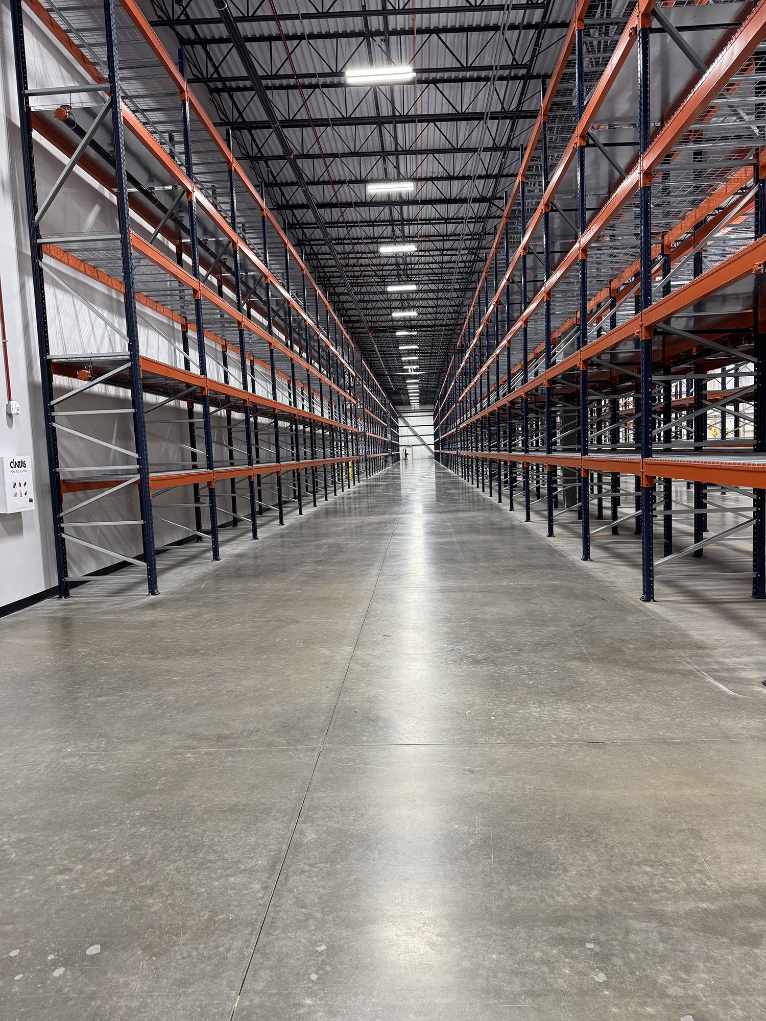 Empty warehouse aisle with tall orange and blue metal shelving units on either side, gray concrete floor, and ceiling with exposed beams and fluorescent lights.