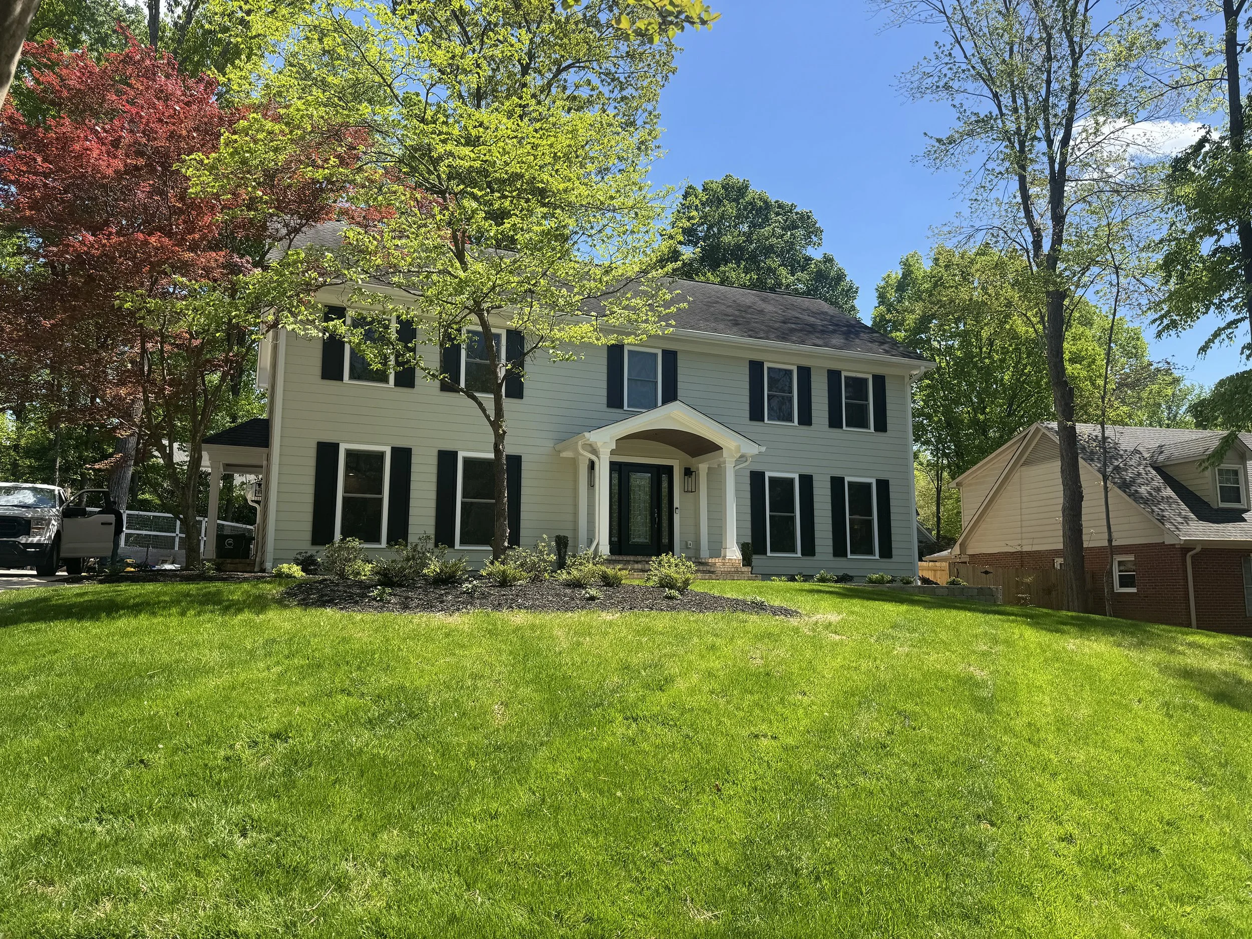 Front yard of a house with green grass, two trees, and a driveway with a white pickup truck. The house is two stories with white siding, black shutters, and a covered front porch. The sky is clear and blue.
