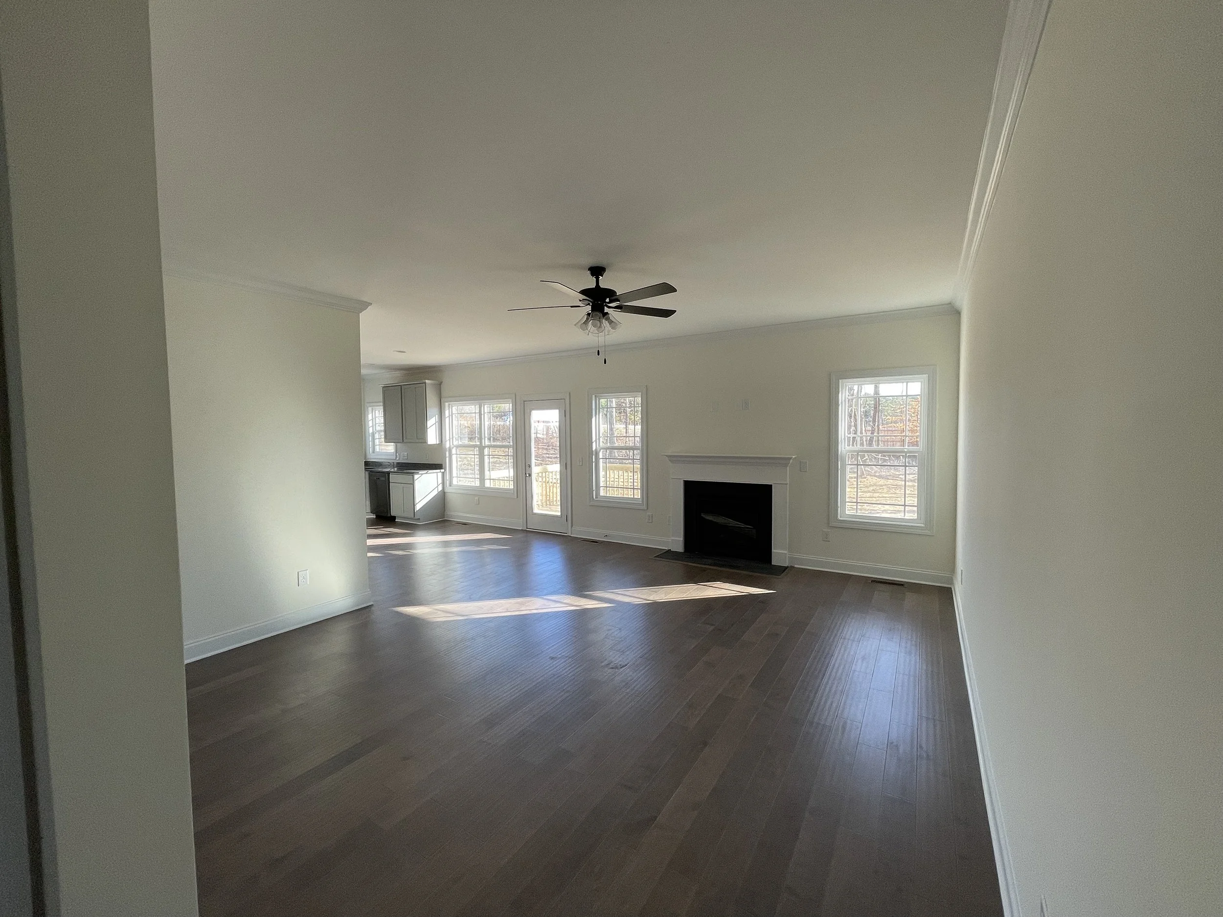 A spacious living room with hardwood floors, white walls, large windows letting in natural light, a ceiling fan, and a fireplace.